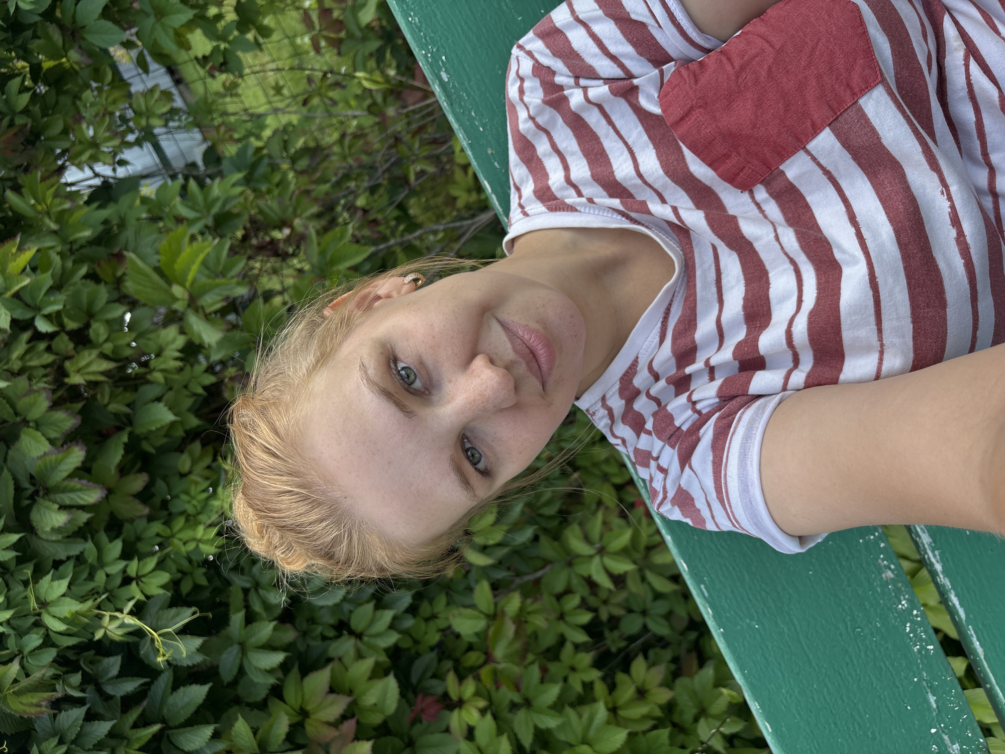 A young Caucasian woman with blonde hair pulled into a bun and freckles on her face is pictured from the chest up, looking directly at the camera. She is wearing a white t-shirt with red stripes and a solid red pocket on the left side. She has light green eyes and is wearing a small hoop earring in her left ear. The woman is sitting on a green wooden bench, which is partially visible in the foreground. Behind her, there is a dense wall of green foliage, which appears to be a hedge or bushes, and a glimpse of a fence and some structures in the background. The lighting suggests it is daytime, possibly overcast, as there are no harsh shadows. The image does not contain any specific identifying features of the city of Дунилово, Russia.