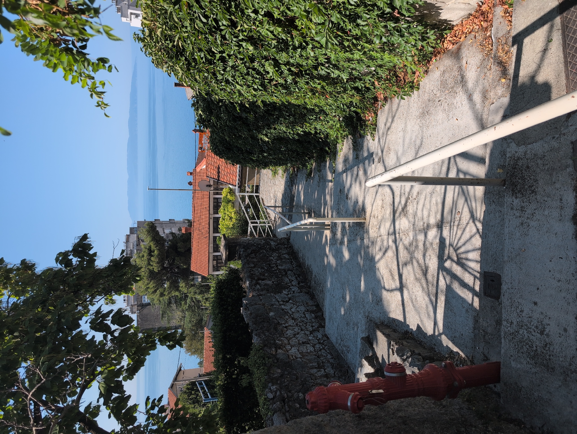 This is a vertical, eye-level, medium shot of a concrete pathway leading downhill towards a body of water, likely the Adriatic Sea, given the location context of Grad Rijeka, Croatia. The pathway is bordered by lush green foliage on the left and a white metal railing on the right. Sunlight casts sharp, angular shadows from an unseen structure onto the concrete, indicating a sunny day.
In the foreground, a bright red fire hydrant is visible on the right side of the pathway. Further down the path, stone walls and terracotta-roofed buildings are partially obscured by trees and more greenery. The distant landscape features buildings and hints of mountainous terrain across the expanse of blue water, under a clear blue sky.
The scene appears to be a quiet, residential area with a view of the sea. The time of day is likely late morning or early afternoon, judging by the length and angle of the shadows. There is no visible text in the image. The overall mood is one of tranquility and pleasant weather.
