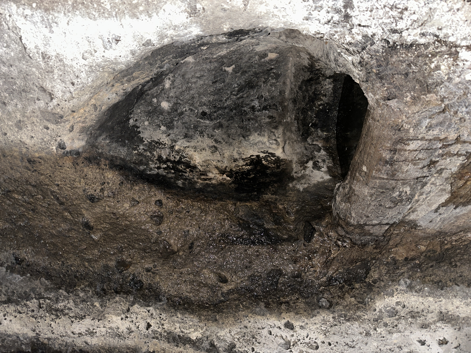 This image displays a close-up view of a damp, textured rock formation in Goght, Armenia. The scene is dominated by an irregular, rough stone surface mottled with shades of grey, brown, and black, punctuated by lighter, almost white patches of weathered rock or mineral deposits along the left and right margins. A prominent, dark, tear-drop shaped cavity or recess is visible in the upper-right section of the frame. Below this, a smaller, horizontal opening is present at the bottom-right, partially filled with a pool of dark liquid, consistent with stagnant water. A rough, horizontally oriented stone projection extends from the left into this lower opening, appearing as a natural channel or ledge. The central area of the rock face exhibits numerous glistening wet spots and dark vertical streaks, indicative of active water seepage or previous flow. The surface texture is granular and uneven, with small embedded mineral fragments visible across the damp areas. No people or animals are present.