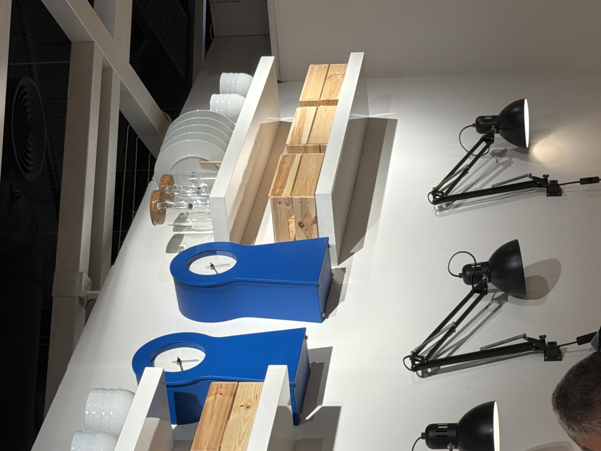 In Varna, Bulgaria, this image depicts an indoor scene with two modern, bright blue grandfather clocks mounted on a white wall. Each clock has a circular face with a black hand. Below the clocks, white shelves display various items. The upper shelf holds stack of white plates and two white cups. The lower shelf contains several small wooden crates. In the foreground, three black adjustable desk lamps are mounted on the wall, casting light downwards. One lamp is closer to the left edge, another is in the center and the third is towards the right edge of the image, all are positioned above the viewer's eye level. A person's head and shoulders are partially visible at the bottom left of the frame, looking upwards. The ceiling features a black ventilation grate and light-colored structural beams.