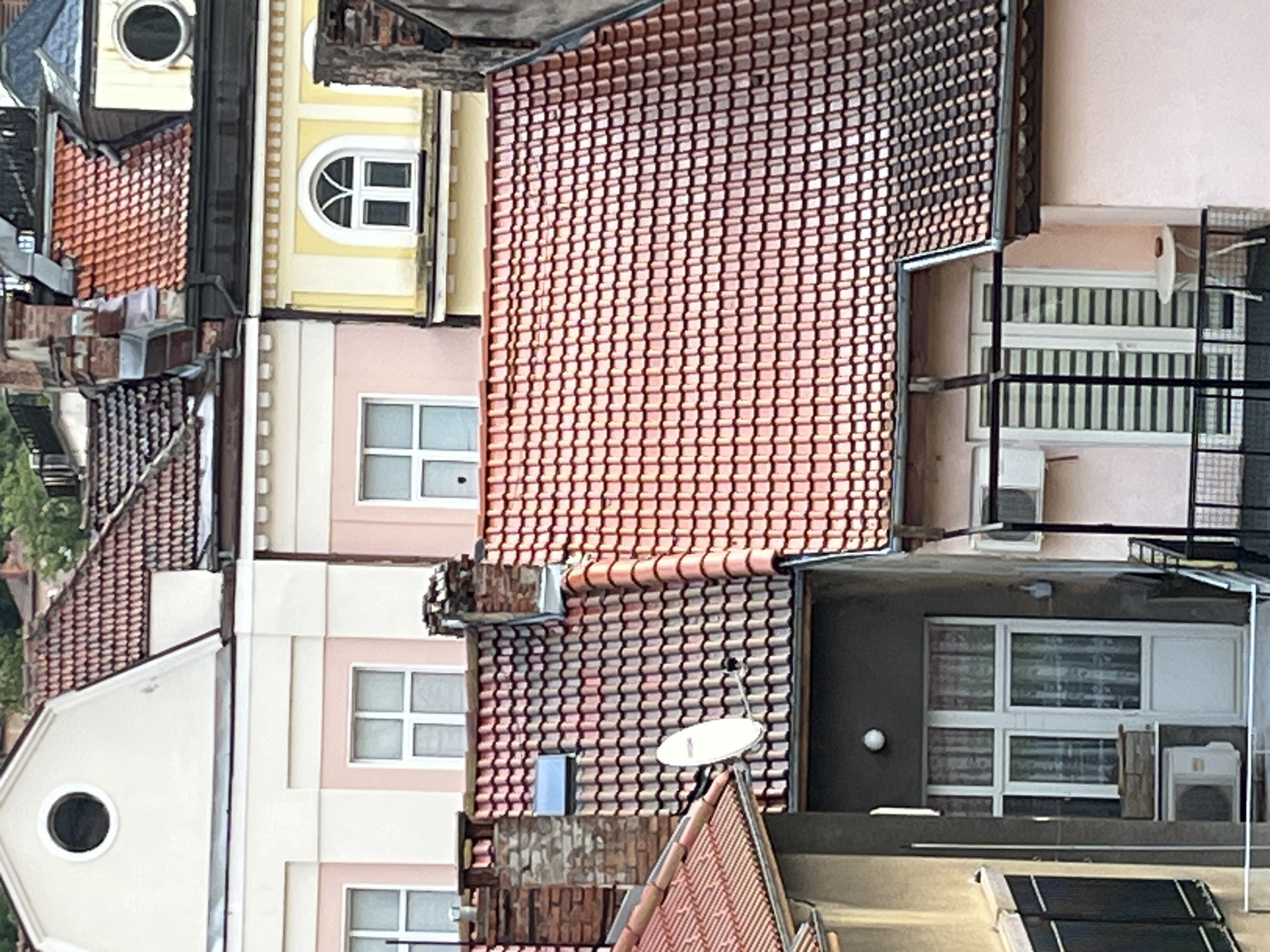 The image provides a high-angle view looking down upon a cluster of interconnected buildings, presenting a densely packed urban or semi-urban landscape. The scene appears to be rotated 90 degrees clockwise.
The primary subjects are the roofs and upper facades of multiple residential or commercial structures. The majority of the roofs are covered with reddish-brown or terracotta-colored tiles, arranged in a regular, interlocking pattern. Several building facades are visible, painted in light hues such as pale yellow, light pink, and beige, often with white trim around windows.
Various types of windows dot the buildings, including rectangular windows with white frames, some showing blinds or curtains inside, an arched window with mullions, and a circular porthole window. On some ledges and walls, outdoor air conditioning (HVAC) units are visible. A small white satellite dish is mounted on one of the tiled roofs. On the lowest visible rooftop, several black rectangular panels, likely solar panels, are installed. Metal railings and grates are also noticeable on some balconies or window areas.
There are no people visible, and no specific activity is taking place. The scene is static, captured during daytime under what appears to be overcast or diffused lighting conditions, as shadows are soft and muted. There is no discernible text in the image. The architectural styles, particularly the tiled roofs and window designs, suggest a European setting.