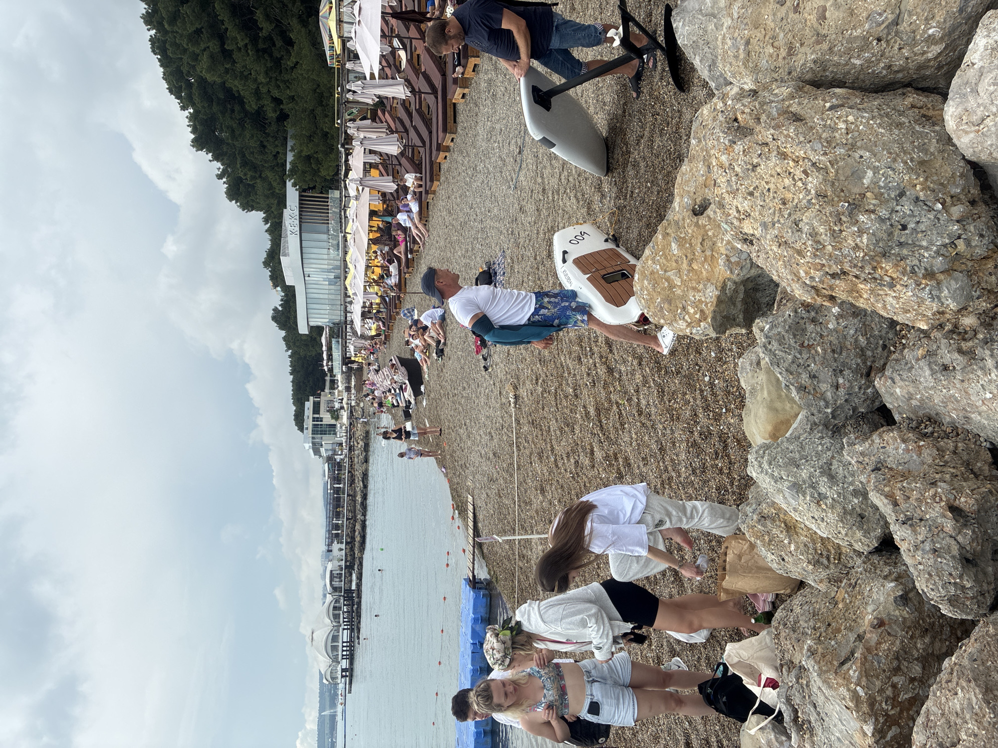 The image depicts a pebble beach in Gelendzhik, Russia, with the Black Sea in the background. Several people are scattered across the beach, some relaxing on lounge chairs under umbrellas, while others are standing or walking. In the foreground, a group of young adults are gathered. Two men are seen near two watercrafts, one appearing to be a hydrofoil. The water is calm, and a few individuals are swimming. In the distance, a modern building with a large white domed roof and a sign that reads "KEKC" is visible, suggesting it might be a restaurant or entertainment venue. The sky is overcast, indicating a cloudy day.