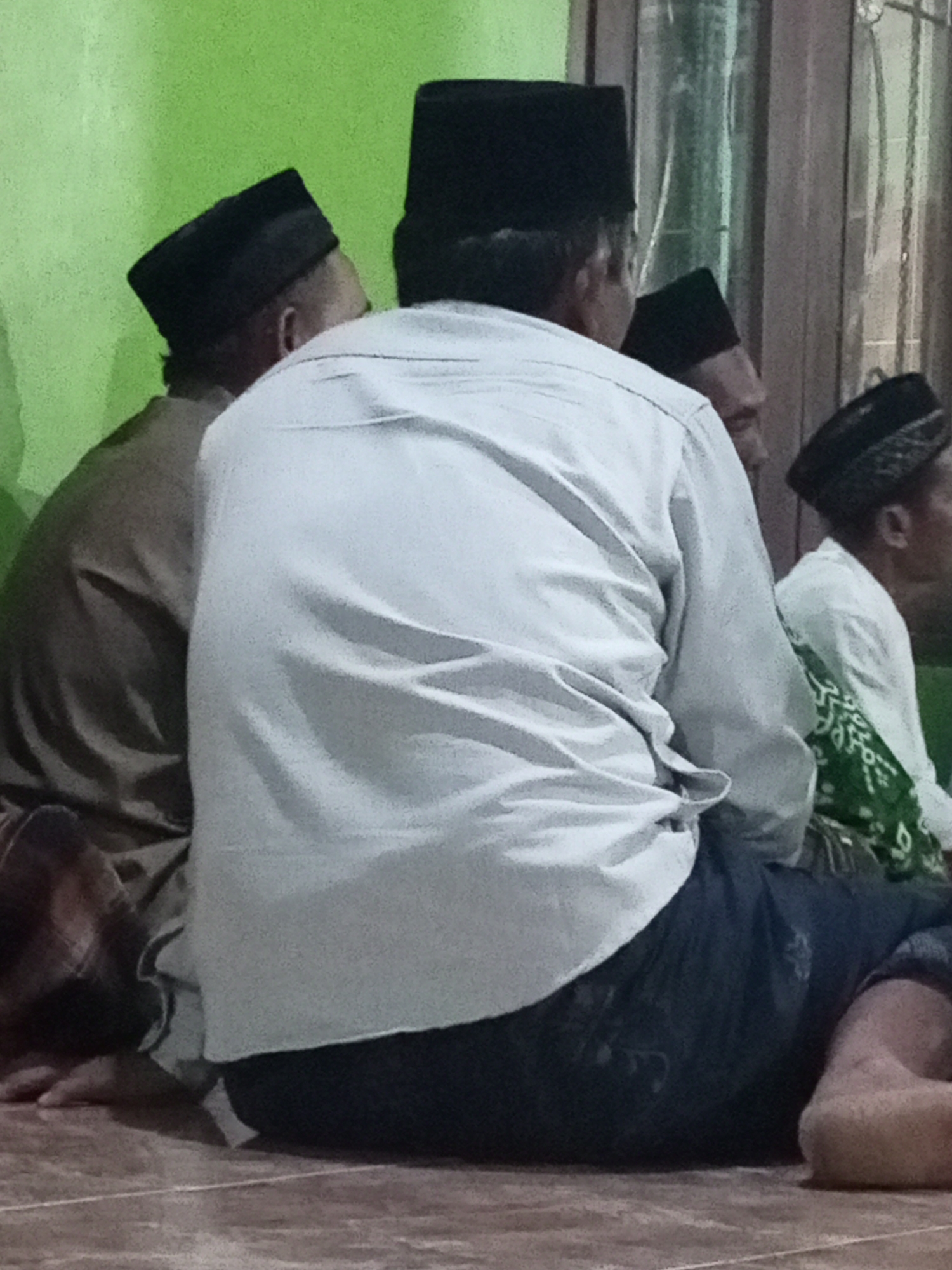 In the city of Lemahbang, Indonesia, four individuals are seated on the floor, seemingly in a religious gathering or ceremony. The person in the foreground, viewed from the back, is wearing a light-colored long-sleeved shirt and dark pants, along with a black, rectangular prayer cap. To their left, another individual, also wearing a black prayer cap, is visible from the side, dressed in a brown garment. Further to the right, two more individuals are present. One is wearing a white shirt and a black prayer cap adorned with a patterned band, and their lower body is partially obscured by a green and white patterned fabric. The fourth individual is partially visible to the right, also wearing a black prayer cap. The environment appears to be indoors, with a green wall on the left and a wooden door or window frame on the right. The floor is tiled with a brownish-red hue.