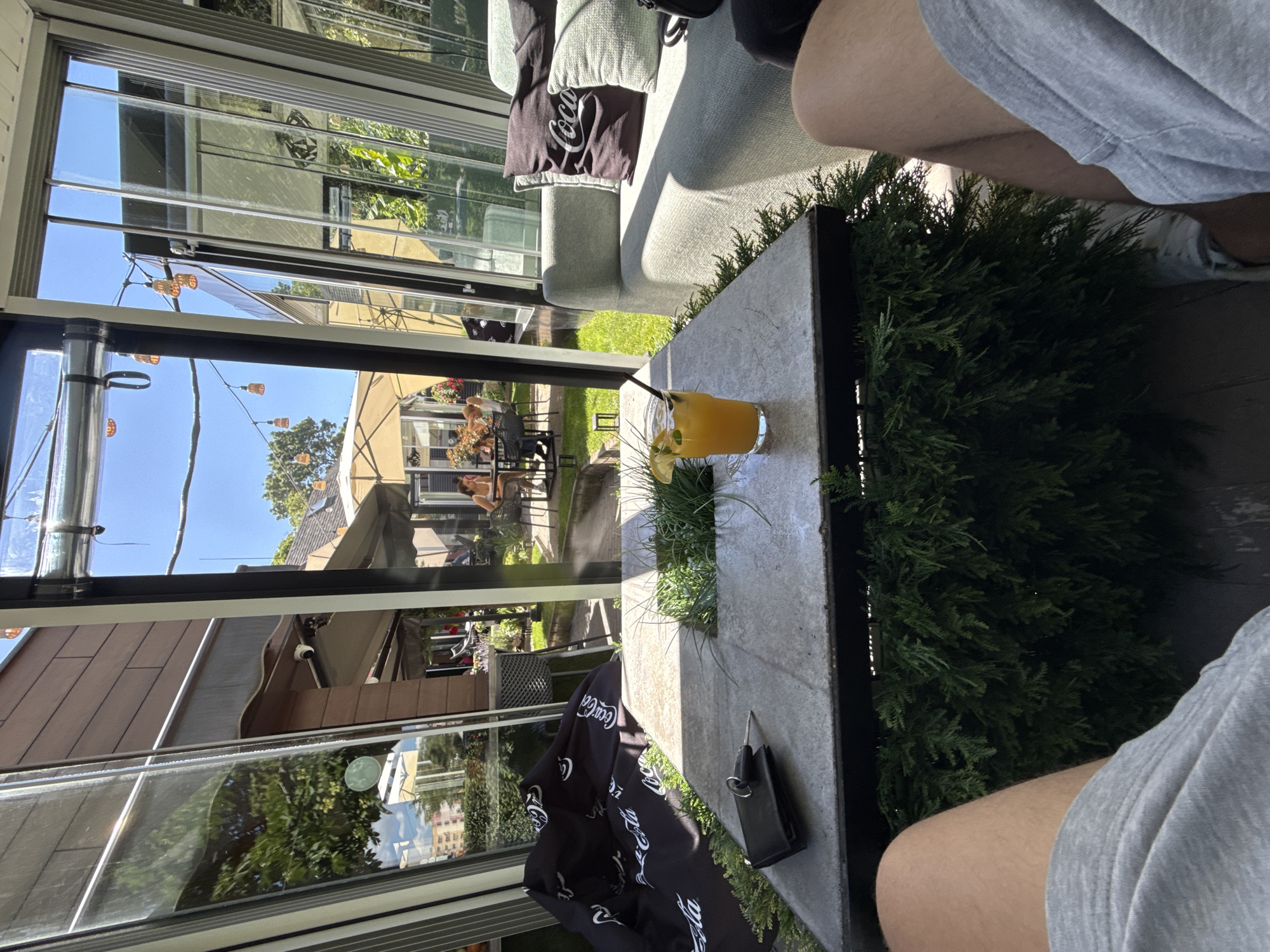 The image shows a perspective from a person sitting in a glass-enclosed area, looking outwards. In the foreground, a square-shaped table with a stone-like surface is visible, topped with a glass containing an orange beverage with a straw and a lemon wedge. To the left of the drink, a black wallet and keys are placed on the table. The table itself is surrounded by dense green foliage. Beyond the table, a light-colored couch with pillows is present.
The glass enclosure opens to an outdoor area with a green lawn and trees. Several people are seated at tables in the background, some under umbrellas. String lights are visible hanging overhead. The architecture suggests a cafe or restaurant with an outdoor seating area. The presence of a Coca-Cola branded beanbag further indicates a commercial establishment. The overall scene depicts a relaxed outdoor setting on a sunny day. The location is specified as Kyustendil, Bulgaria.