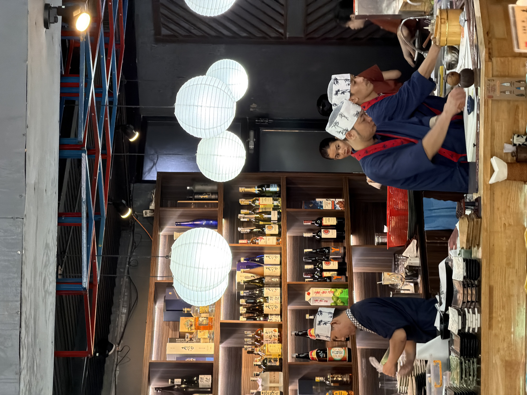 The image depicts the interior of a sushi restaurant in Nha Trang Ward, Vietnam. The restaurant features wooden shelves stocked with various bottles of alcohol, including sake and wine. Several white paper lanterns hang from the ceiling, providing illumination. In the foreground, three men in blue chef uniforms with white patterned headwear are working behind a wooden counter. One man is preparing food, while another is engaged in a task at the counter. A woman is visible in the background, looking towards the chefs. Stacks of plates and bowls are neatly arranged on the counter. The overall ambiance suggests a busy and functional restaurant setting.