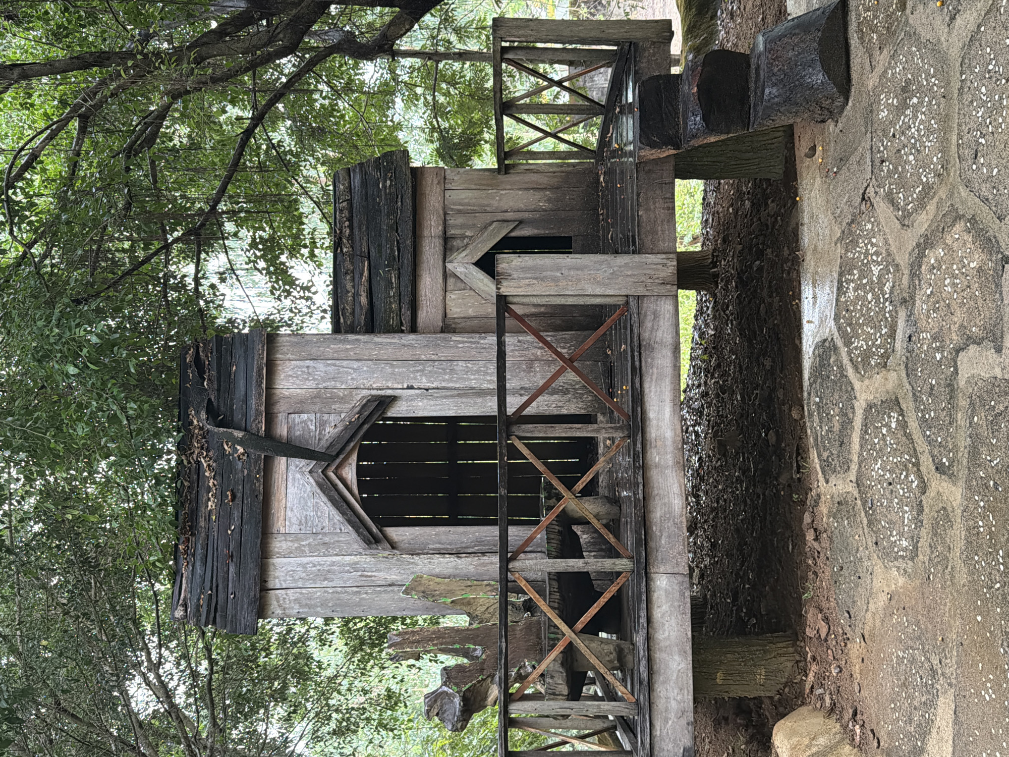 This image depicts a wooden structure in Khánh Vĩnh, Vietnam, possibly a small building or playhouse, seemingly situated outdoors within a wooded area. The structure has two stories and an irregular design, exhibiting features such as a ladder, railing, and window openings, some of which are decoratively shaped. The building shows evidence of aging and weathering. The surrounding environment contains trees, foliage, and a paved pathway, suggesting a park or recreational area. The image appears to be a photograph taken in natural light.