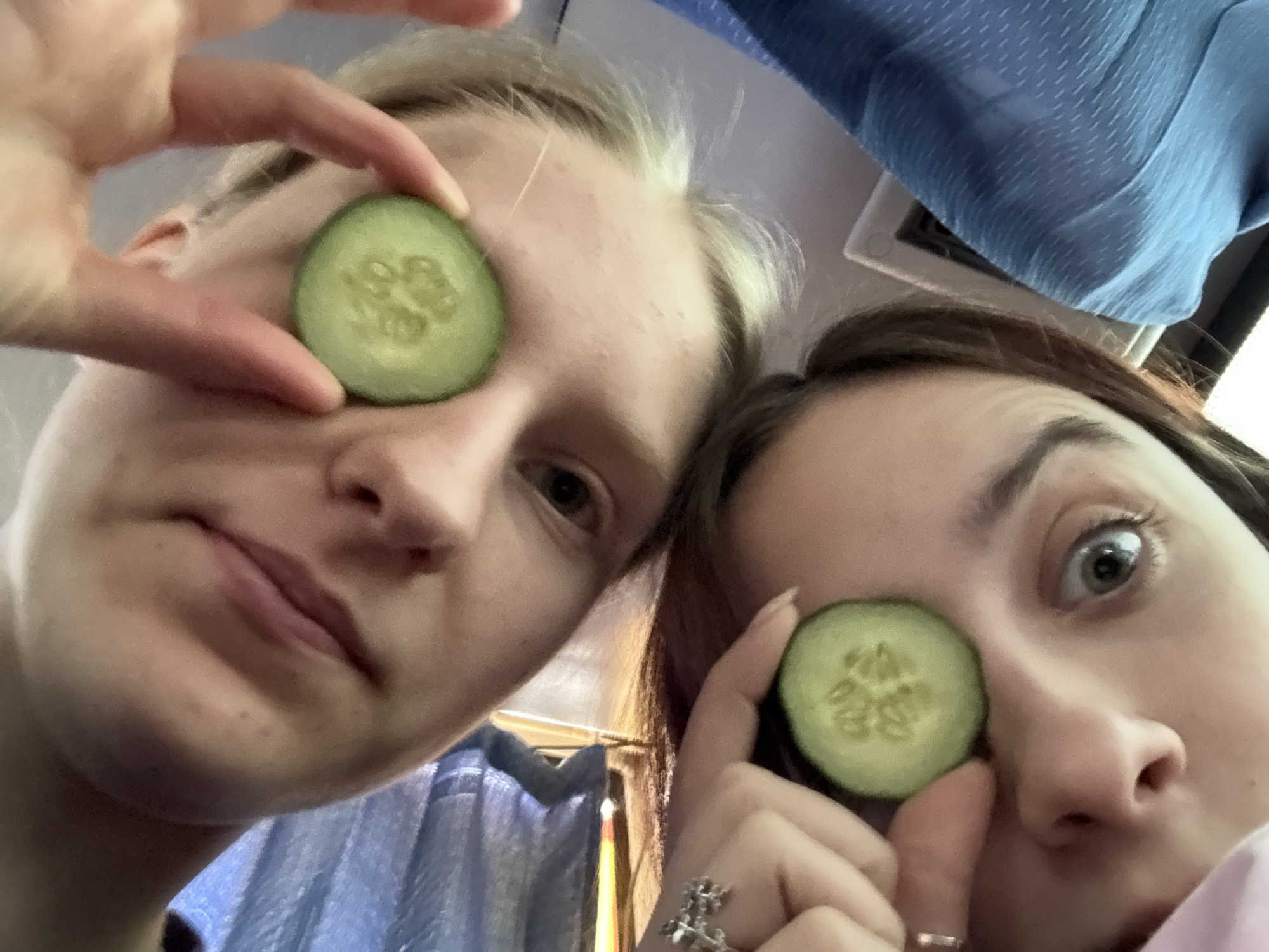 Two individuals are visible in the photograph, each holding a cucumber slice over one eye. The person on the left has fair skin and blond hair. The person on the right has brown hair. The background includes draped blue fabric. The location of Yanychi, Russia, is not discernible from the visual information provided.