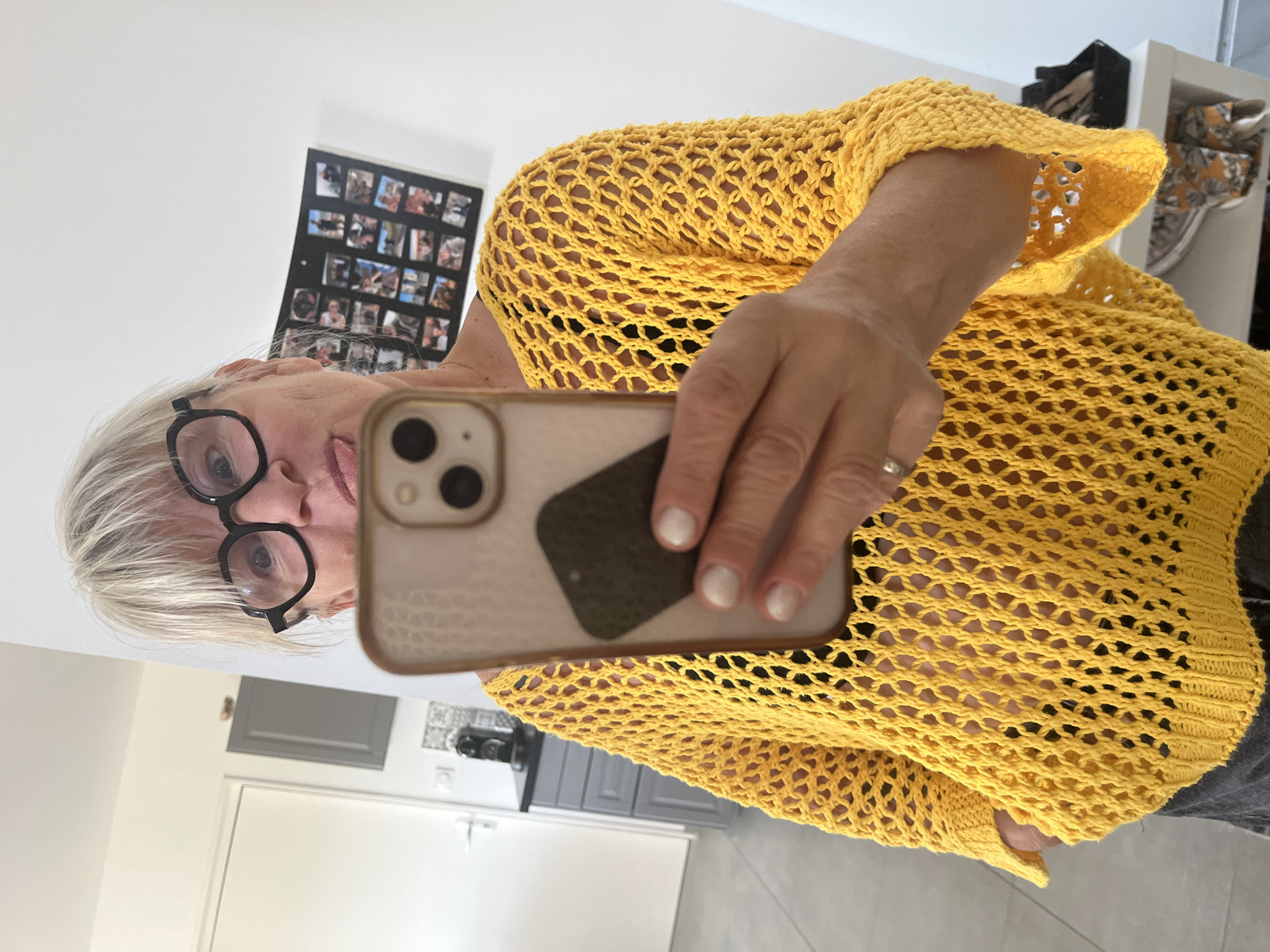 A woman with blonde hair and black-rimmed glasses is taking a selfie in a room that appears to be in a home. She is wearing a yellow, open-knit sweater over a dark bra. She has her tongue sticking out slightly. In the background, there is a door, kitchen cabinets, and a framed collage of photographs on the wall. The scene is lit by artificial light. The location is not specified as Chambœuf, France.