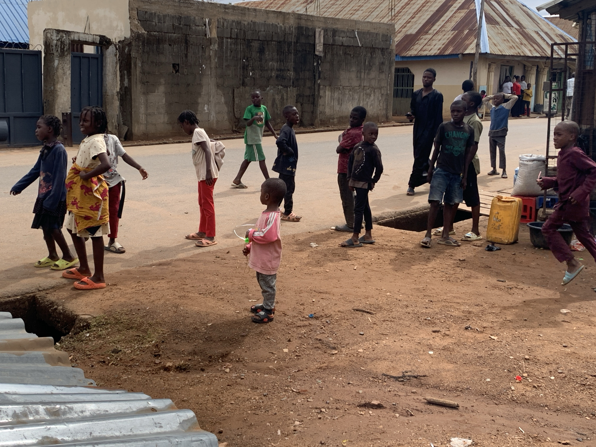 The photograph captures a group of children in Akwanga, Nigeria, standing near a dirt road beside a concrete building. The children are of various ages and attire, some wearing sandals or flip-flops. Some children are walking along the road, others are standing and facing forward, and one is running. The road is unpaved, with a narrow strip of concrete serving as a sidewalk in sections. Several other buildings are visible in the background, including one with a corrugated iron roof. An open drain is partially visible in the foreground. The setting suggests a residential or commercial area in a developing urban landscape.
