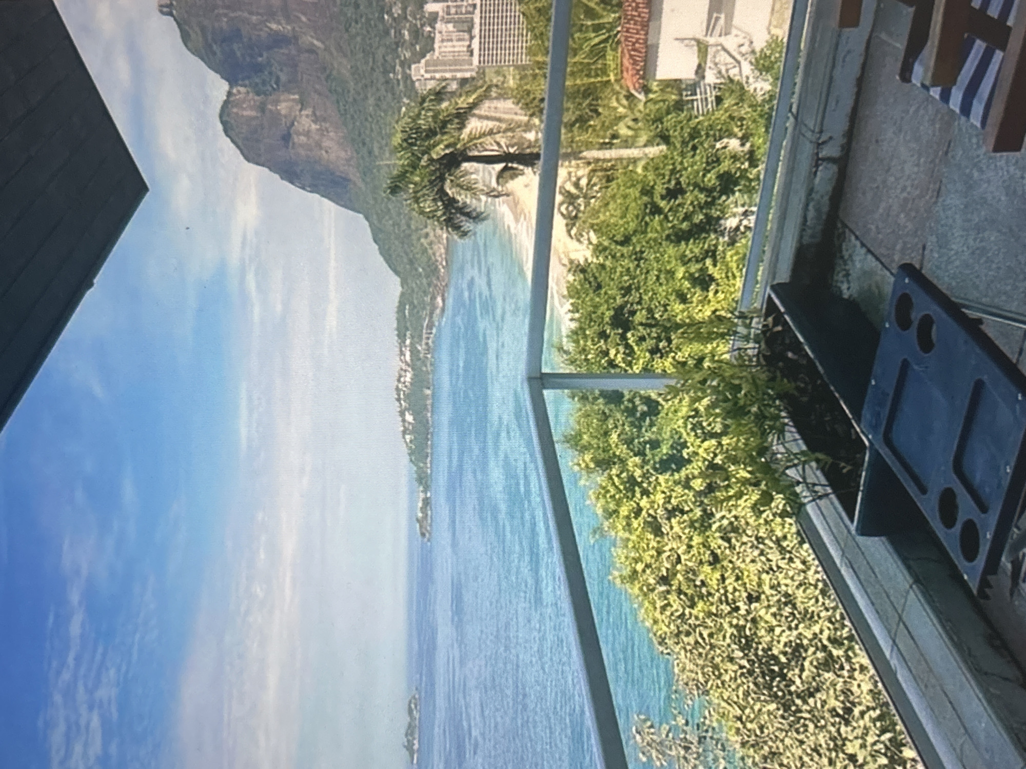 The image depicts a scenic view of a coastline with a mountain in the background. The ocean is a vibrant blue, meeting a sandy beach that is lined with lush green trees and some buildings. A palm tree is prominently featured in the mid-ground. In the foreground, a balcony railing is visible, suggesting the vantage point is from a high-rise or a terrace. A dark blue portable grill with black circular indentations is placed on the balcony floor, with striped cushions of a chair visible to its right. The sky is a clear blue with scattered white clouds, indicating it is daytime. The overall impression is one of a beautiful, sunny day in a coastal resort area. There is no visible text in the image.