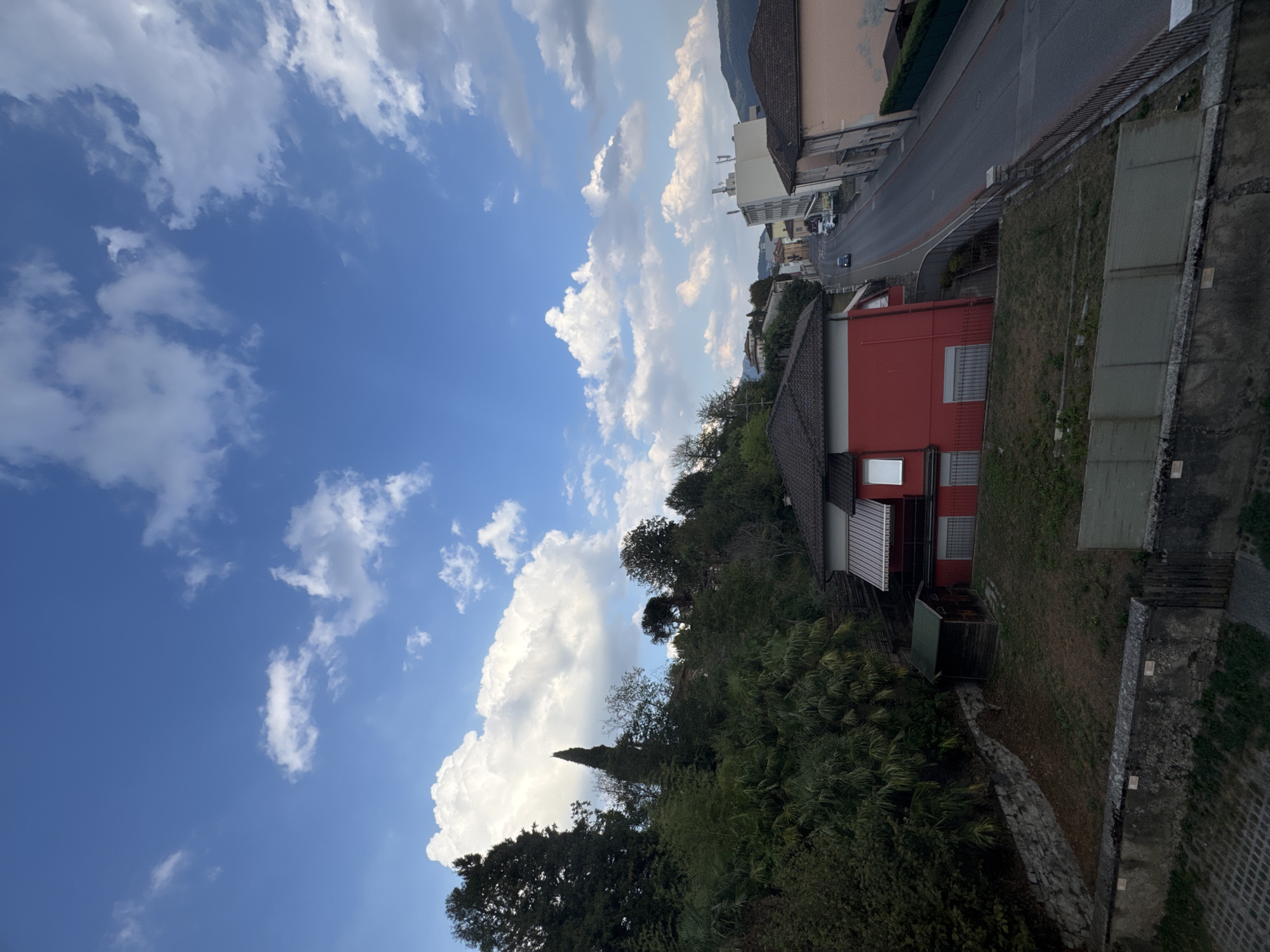 The image shows a view of a residential area in Balerna, Switzerland. The foreground features a concrete wall and a grey metal fence. Behind the fence is a grassy area with some dry patches, bordered by a low stone wall. To the left, a steep embankment is covered in dense green foliage and trees.
In the middle ground, a bright red house with a dark tiled roof is prominent. The house has white-trimmed windows and an awning over a balcony. Adjacent to the red house is a small, dark wooden shed with a green roof.
The background shows a street winding uphill, lined with buildings of varying heights and architectural styles. One building is a tall, modern white structure. Further up the street, more houses and trees are visible, with distant hills forming the horizon. The sky above is a clear blue, dotted with white, fluffy cumulus clouds. A few cars are visible on the street. The overall impression is a daytime scene in a European town.