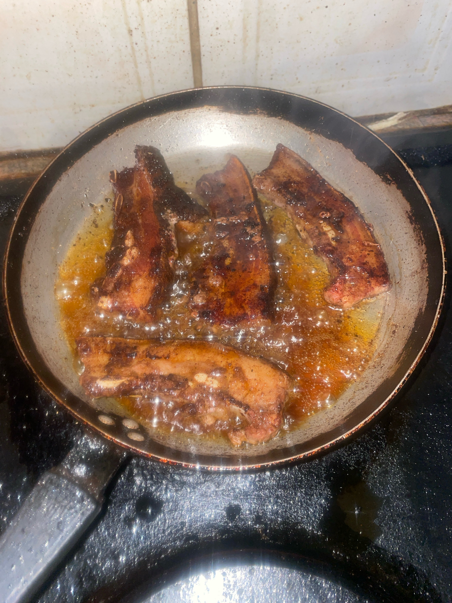 In Bronkhorstspruit, South Africa, four pieces of seasoned meat are sizzling in a frying pan. The pan is on a black stovetop, with white tiled walls in the background. The meat is browned and appears to be pork belly, rendered fat is bubbling around it. The lighting is dim, suggesting an indoor setting at night or with low light.