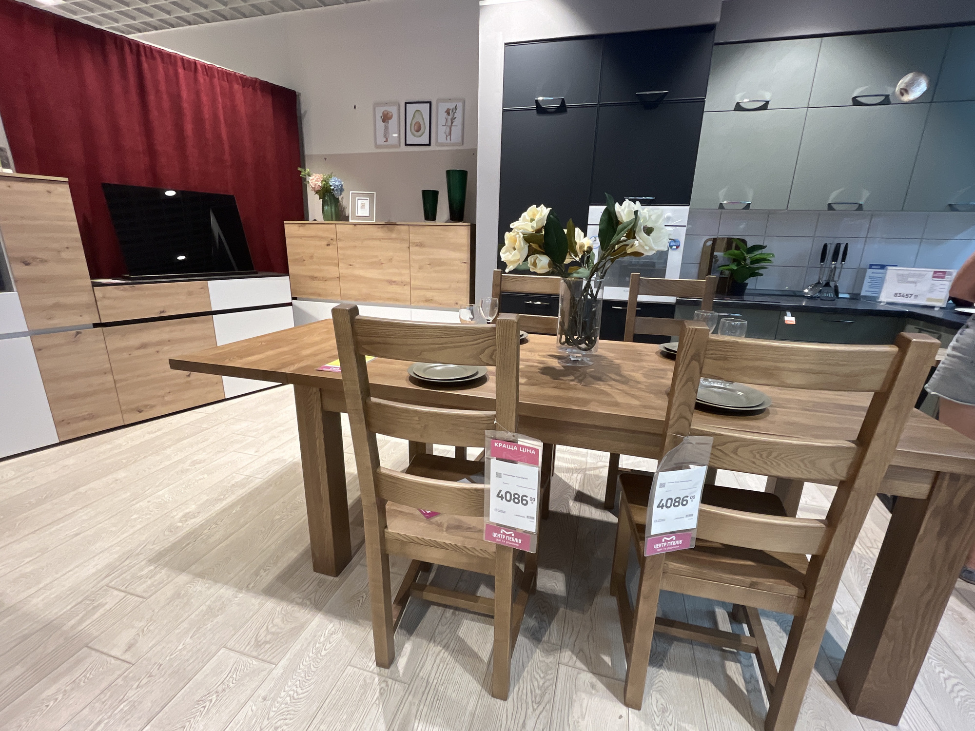 The image shows a dining set in a furniture store. The dining set includes a wooden table and four wooden chairs. The chairs have tags attached to them that display prices. In the background, there is a wall with red curtains, a TV, and kitchen cabinets. The floor is made of light-colored wood planks. The lighting is bright and appears to be artificial, typical of a retail environment. The scene is static and does not show any people actively engaged in an activity.