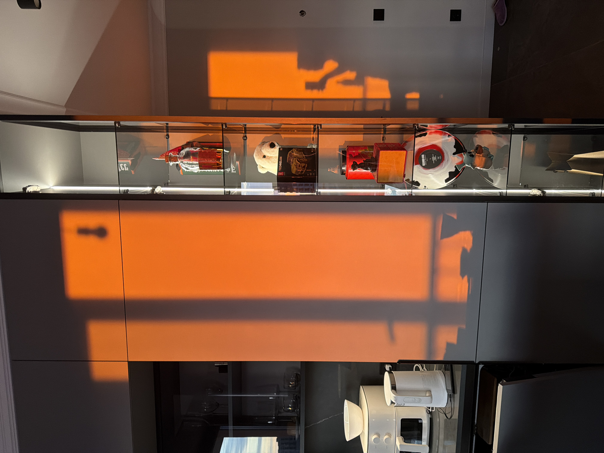 The image displays a modern interior space, likely a kitchen or living area, illuminated by intense, low-angle orange sunlight, suggestive of either sunrise or sunset. The specific location within the city of Minsk, Belarus, cannot be determined from the visual evidence provided.
On the left, a section of dark grey, handleless kitchen or storage cabinets is visible. A white countertop extends from the left, supporting a white air fryer or similar appliance with a white bowl on top, and a white electric kettle connected via a black cable. A dark smartphone rests on the countertop near the kettle. Behind these, through glass shelving, several clear glass items, including wine glasses and tumblers, are visible. A window on the far left offers a partial view of a sky.
Adjacent to the dark cabinets, centrally positioned, is a tall, narrow display cabinet with internal vertical LED lighting. Within this cabinet, from top to bottom, are: a clear glass bottle with a red stopper containing a reddish liquid; a white plush polar bear and a black box (partially labeled "TECHNOLGY"); a red cylindrical container next to a dark figurine on a red base; a square orange box; a clear vinyl record with a red label ("SECOND SUB") featuring black and white patterns, positioned above a blue figurine; and a brown paper bag-like item on the lowest visible shelf.
The right side of the image features a light-colored wall (off-white or light grey) onto which the strong orange sunlight casts distinct rectangular patterns, indicating window frames. A clear shadow of a person holding a smartphone, capturing the image, is prominently cast on this wall. Two dark electrical outlets or switches are visible on the wall. The floor in the bottom right corner consists of dark wood or laminate planks, with a pair of purple slippers resting on it. The top right reveals a white ceiling with crown molding and a dark, cylindrical ceiling light fixture.