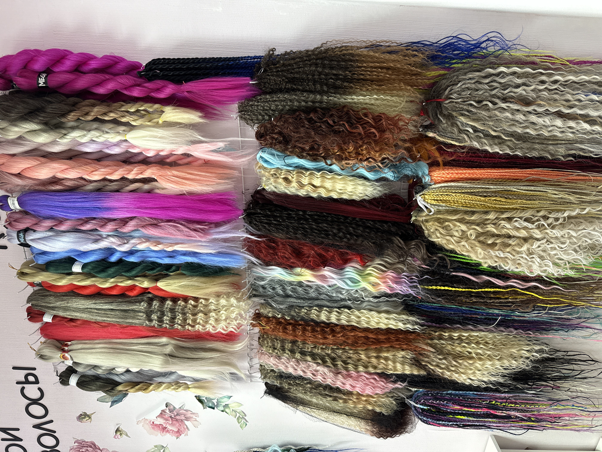 The image displays a comprehensive collection of synthetic hair extensions or braiding hair, arranged on a white vertical surface, likely within a retail or studio environment in Rusanovo, Russia. The scene is static, presenting a colorful inventory of hair products.
The primary subjects are numerous bundles of artificial hair, meticulously organized into three main vertical sections. The color palette is exceptionally diverse, encompassing natural shades of browns, blondes, greys, and black, alongside a vibrant spectrum of artificial hues. These include bright fuchsia, magenta, various purples, blues, greens, reds, peach, and pastel tones. Many bundles showcase multi-tonal blends or ombré effects, with colors seamlessly transitioning along the length of the hair.
Different textures and styles are prominent. The collection includes straight, smooth hair, tightly twisted or braided sections (resembling kanekalon), loose wavy patterns, tight kinky curls, and textured bundles similar to dreadlocks. Many bundles are secured with small white ties, and some are visibly hanging from hooks on the display. A bright pink braided bundle features a white label partially reading "MEGA".
The display area is evenly and brightly lit, characteristic of an indoor setting. Decorative elements include pink rose and green leaf decals adorning the white surface in the bottom left corner. Partially visible Russian text runs vertically along the far left edge, decipherable as "СВОИ ВОЛОСЫ" (SVOI VOLOSY), which translates to "MY HAIR" or "OWN HAIR," suggesting a brand or a product descriptor. The overall presentation indicates a well-stocked and varied selection for hair styling needs.