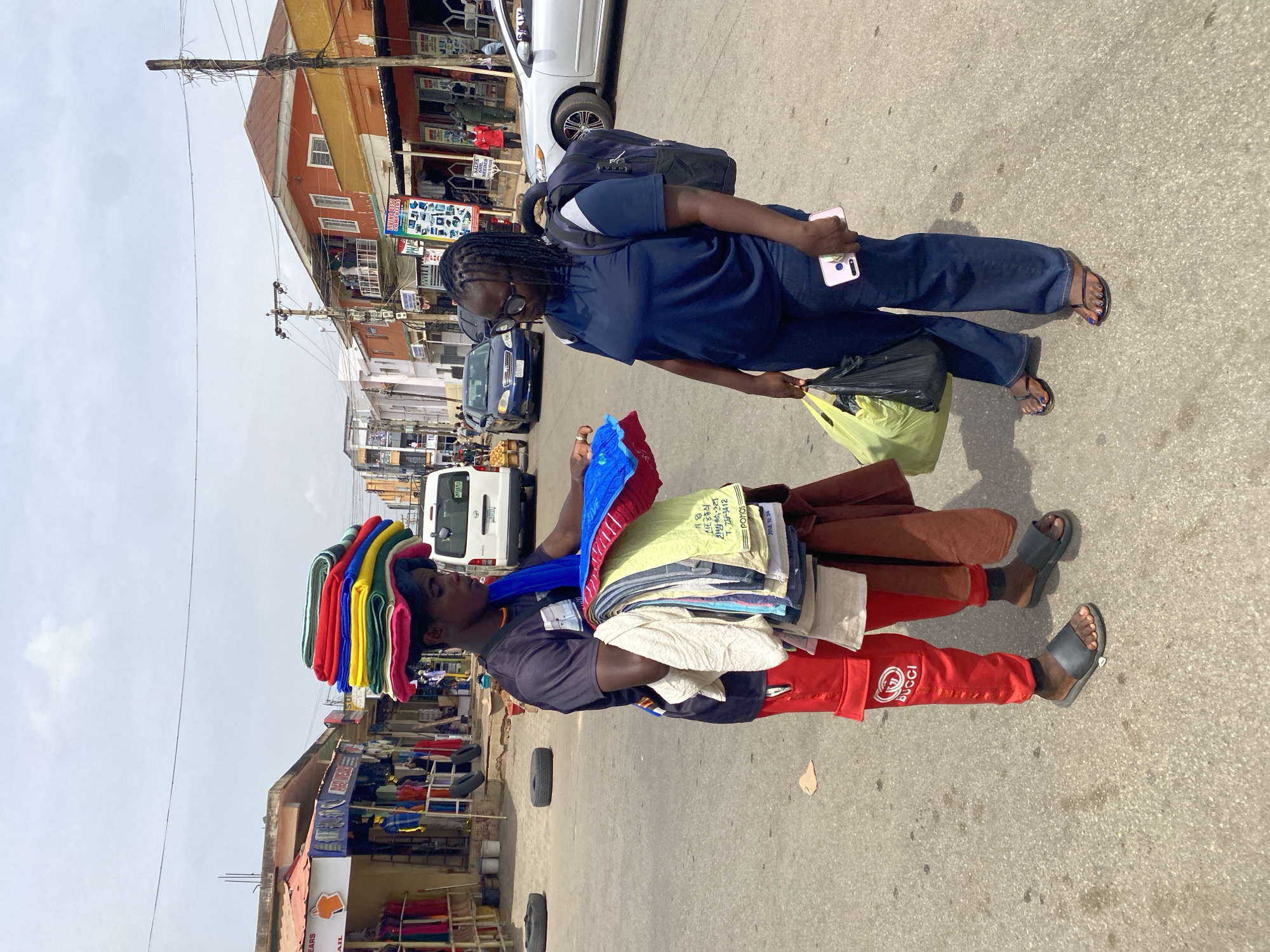 Here's a description of the image:
The image is taken in the city of Jos, Nigeria. Two people stand in a street. The person on the left is carrying a stack of items on their head and is dressed in a dark shirt and red pants with the word "GUCCI" on the side. They are also wearing sandals. The other person, on the right, is wearing a dark blue scrubs-like outfit and a backpack. They are holding a smartphone in one hand and a shopping bag in the other. The background shows shops and buildings along the street, with cars and other vehicles in the distance.
