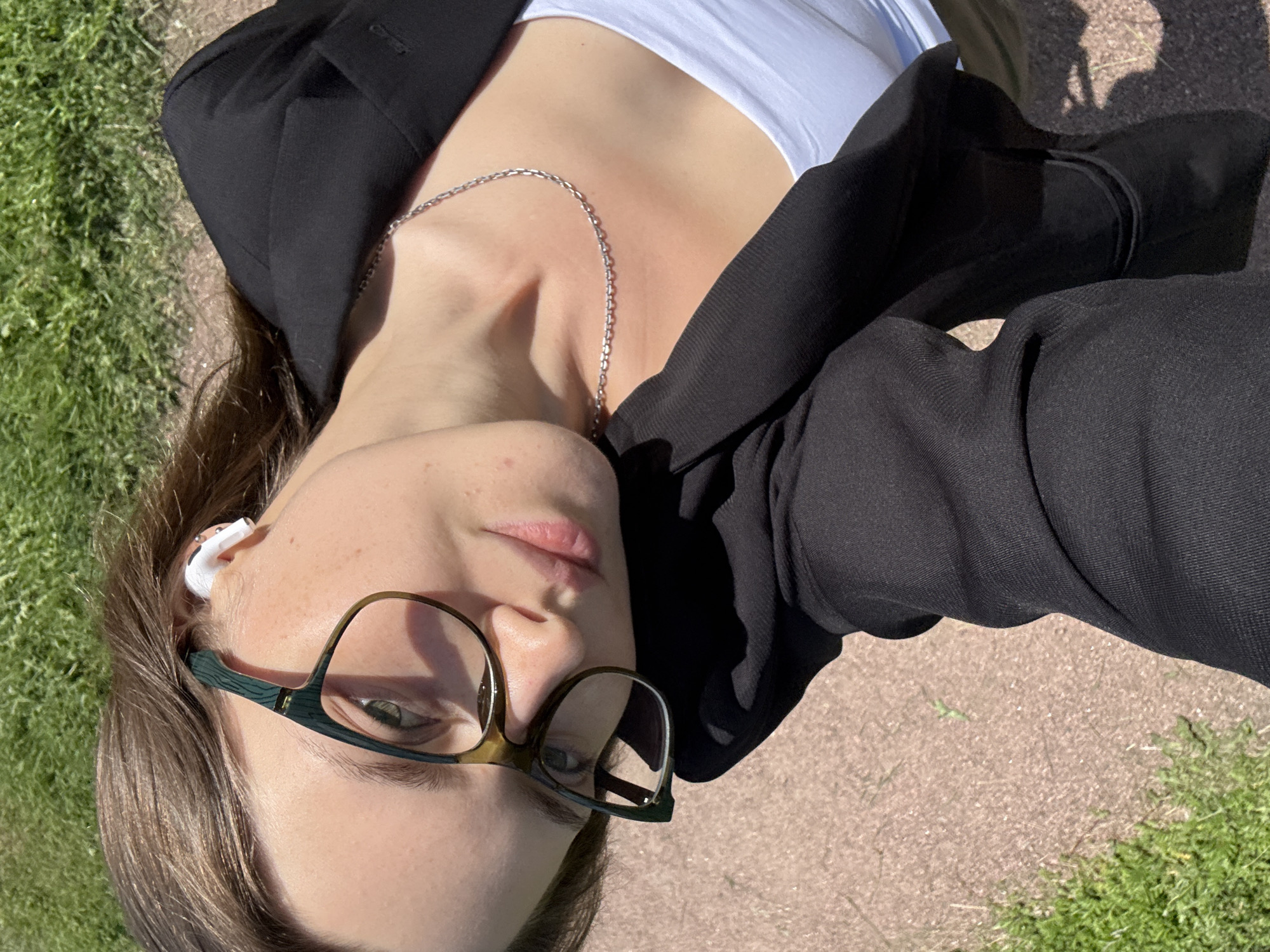 A young woman with brown hair, wearing glasses and earbuds, is captured in a close-up, high-angle shot taken outdoors. She is dressed in a black blazer over a white top and a silver necklace. Her head is tilted, and she is looking towards the camera. The background consists of a pathway with grass and sand, suggesting a park or outdoor area. The lighting indicates it is a sunny day. There is no specific indication of the location being Saint Petersburg, Russia.