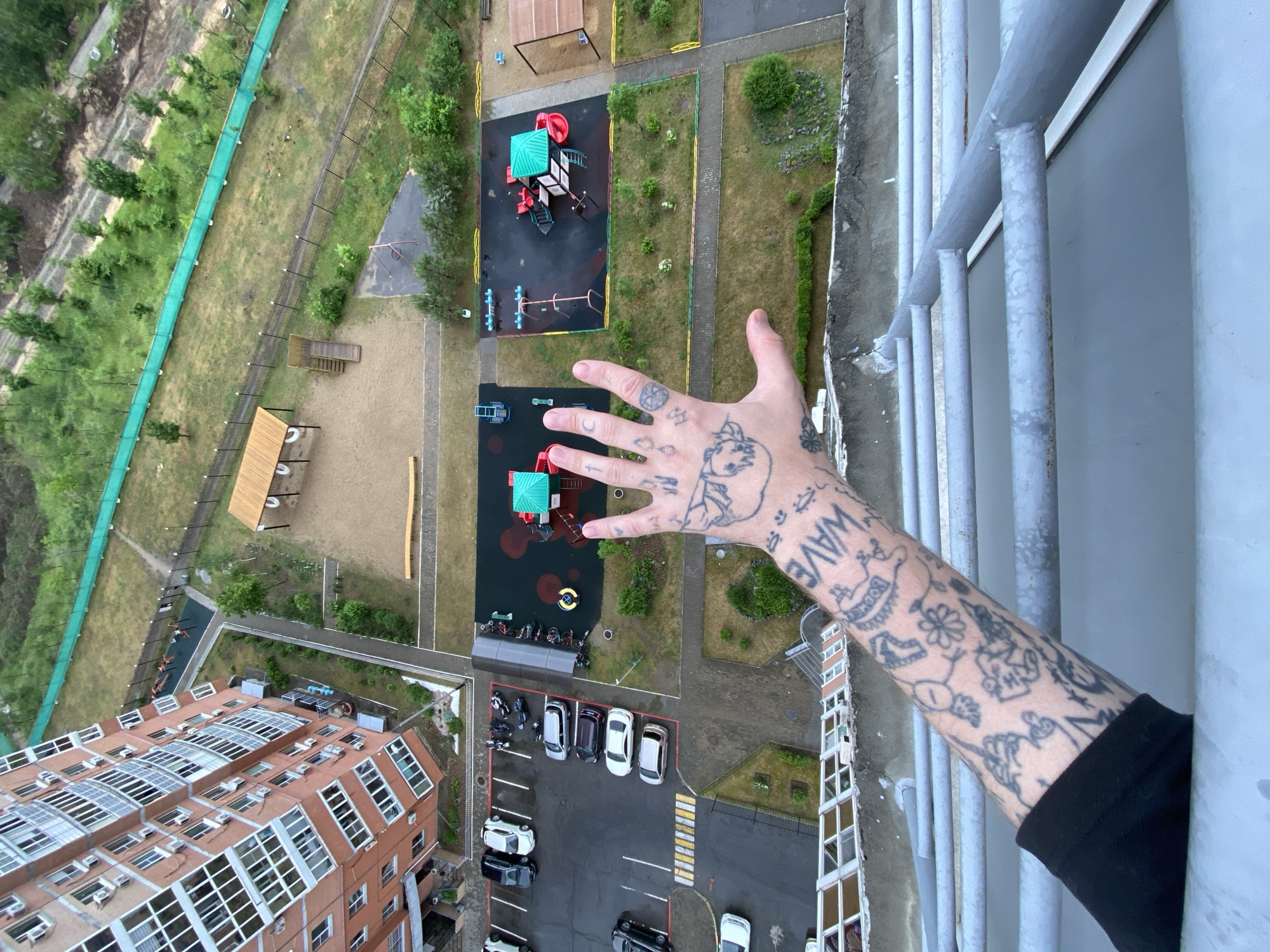The image, taken from a high vantage point in an urban setting, captures a view of a residential building and a playground. The residential building, constructed of brick and featuring numerous windows, dominates the left side of the frame. In the foreground, a person's tattooed arm and hand are visible, extended towards the scene below. The hand displays several tattoos, including what appears to be a crescent moon and a cross. The arm also has numerous other tattoos, some of which are partially obscured by the sleeve of a black garment.
Below the viewpoint, a playground area is visible, equipped with swings, slides, and climbing structures. The ground of the playground is covered with a dark, rubberized surface, with some colorful elements. Adjacent to the playground, a parking lot with several cars and motorcycles is present. Further into the background, there is a grassy area with trees and a green fence.
While the image provides a detailed view of the immediate surroundings, the broader city of Khabarovsk, Russia, is not explicitly identifiable from the visual information alone, beyond the general urban landscape. The scene appears to be a typical view of a residential area with recreational facilities.