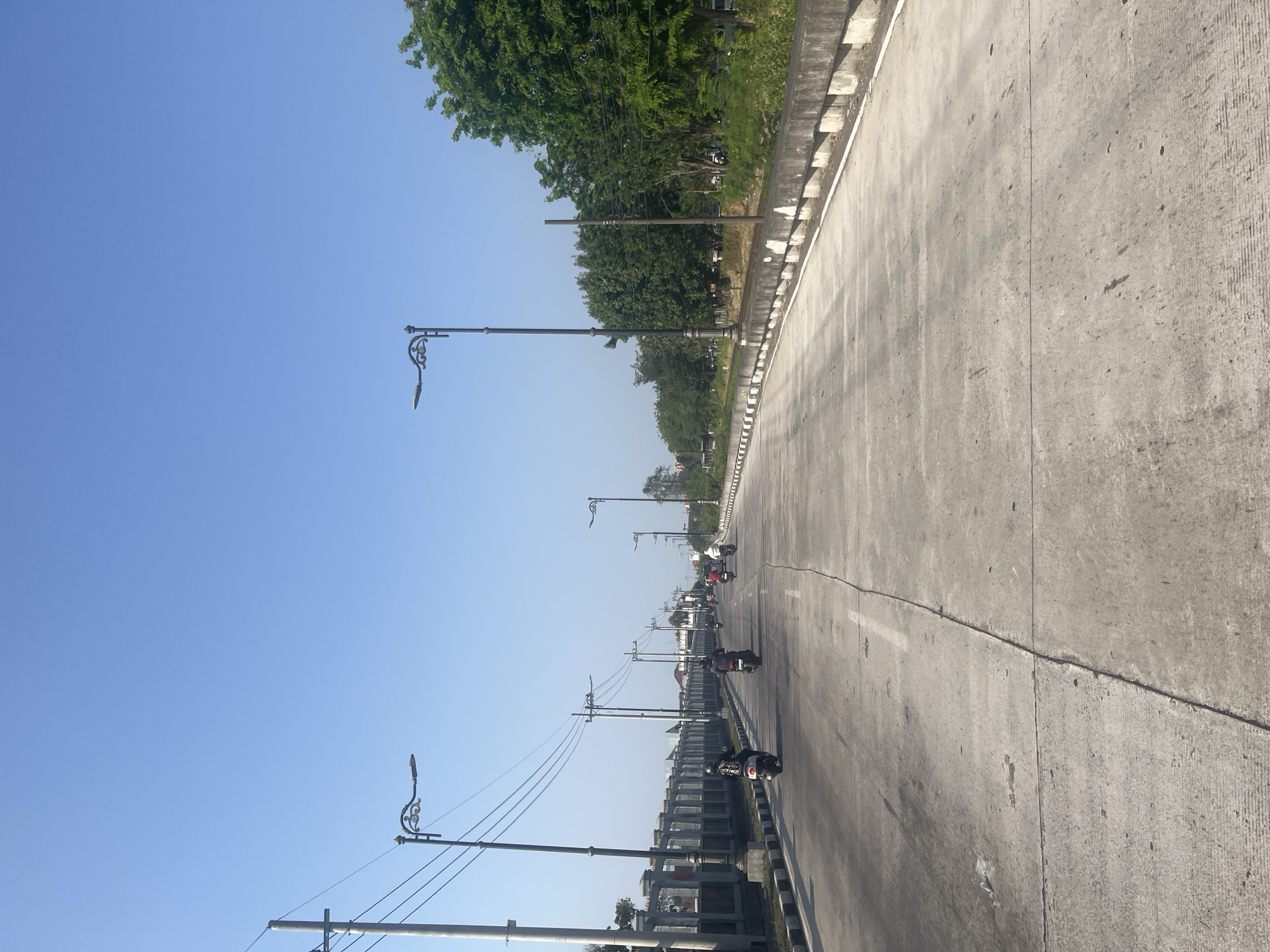 The image shows a road in Gedangan, Indonesia, on a clear day. The road is made of concrete and has multiple lanes. Several motorcycles are seen on the road, with riders wearing helmets. Along the side of the road, there are decorative lampposts and utility poles with overhead wires. A concrete barrier with a black and white striped edge runs along one side of the road, separating it from a grassy area and trees. On the other side of the road, there is a gray concrete wall with decorative posts and railing. The sky is a clear, bright blue.