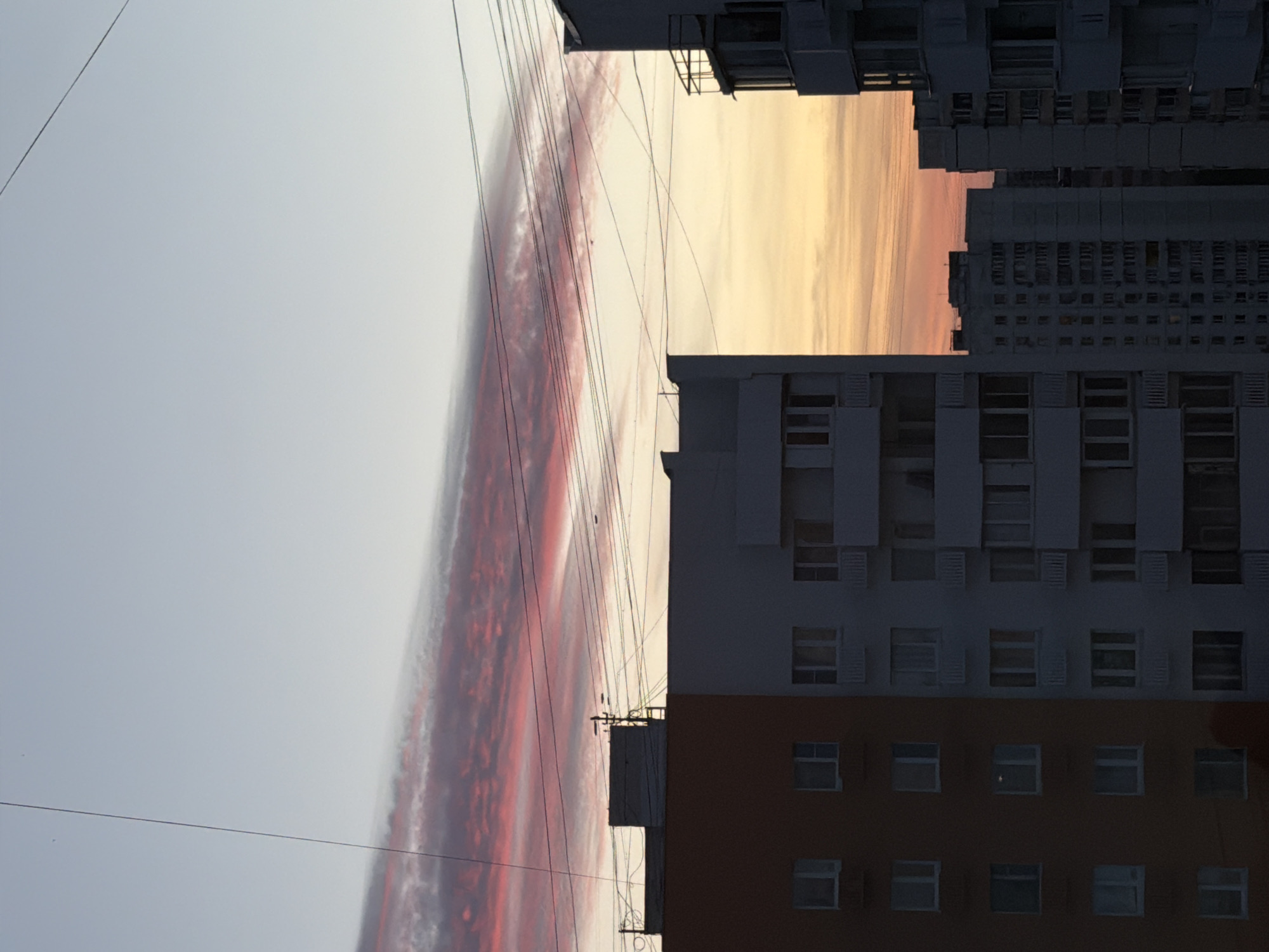 The image captures an urban landscape at either sunrise or sunset, viewed from a low angle looking upwards between towering residential buildings.
Several multi-story apartment blocks dominate the scene. On the right, a prominent building features a reddish-brown lower section and a lighter grey upper part, both adorned with numerous windows, some of which appear to have shutters or blinds. To the left and in the background, additional large, dark grey apartment buildings rise, their facades detailed with many windows and balconies, casting them in shadow against the brighter sky.
The sky is a central element, showcasing a dramatic play of colors. A long, narrow cloud formation stretches horizontally across the middle of the frame, glowing vibrantly with intense reds, oranges, and pinks, illuminated by the low sun. Above this vivid cloud, the sky is a calm, light grey-blue. Below the cloud, between the buildings, the sky emits a warm yellow and orange glow, further emphasizing the dawn or dusk light.
A network of thin, dark wires or power lines crisscrosses the sky, spanning between the buildings and passing over the colorful cloud, highlighting the urban infrastructure. No people or specific activities are visible, allowing the viewer to focus on the architectural forms and the striking atmospheric conditions. The style of the buildings suggests a typical post-Soviet residential area, aligning with the context of Khimki, Russia.