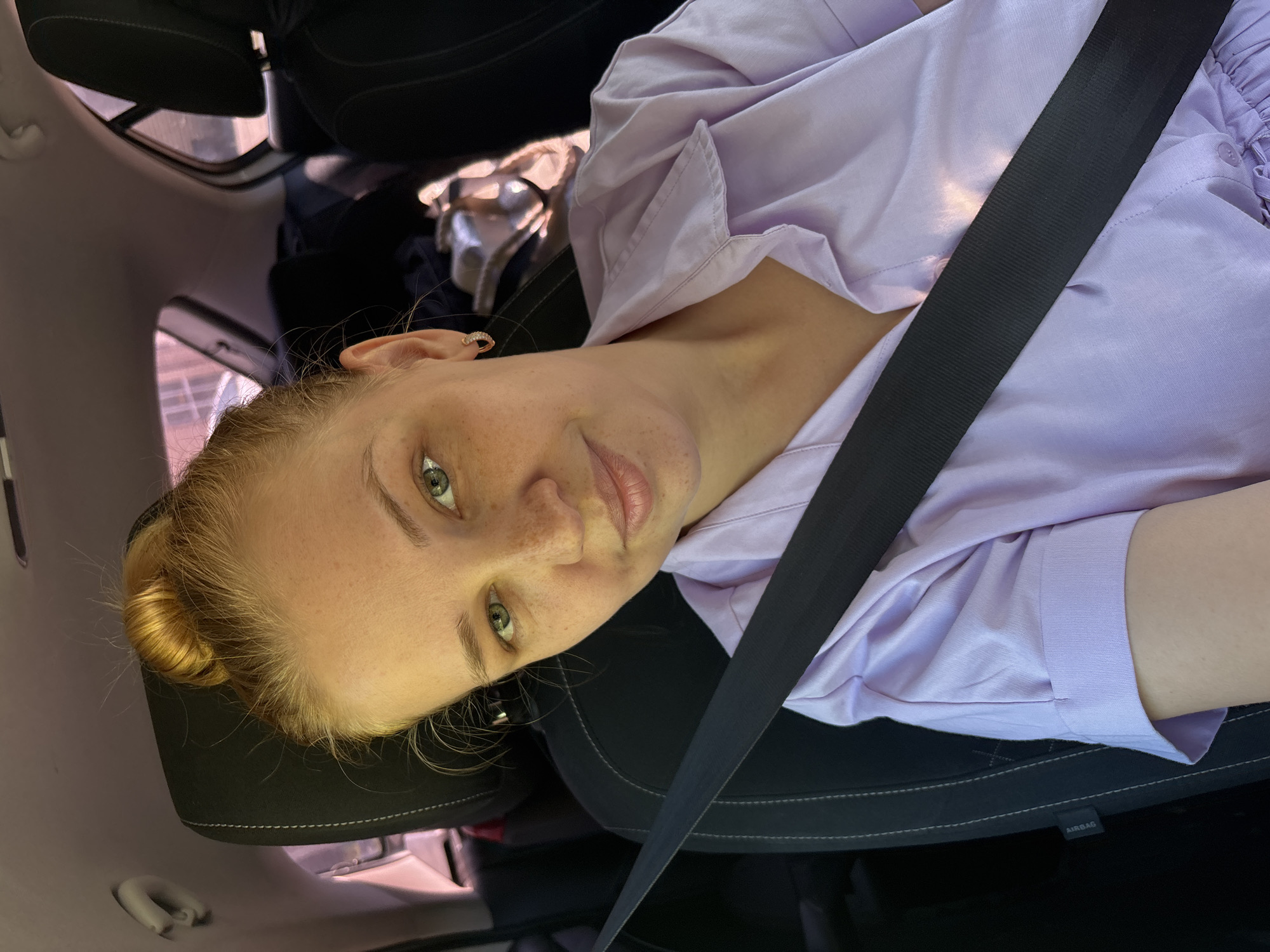 A young Caucasian woman with blonde hair pulled into a bun is seated in the passenger seat of a car. She is wearing a light purple collared shirt and a black seatbelt is across her chest. Her expression is neutral, with her gaze directed towards the camera. The interior of the car is visible, including the headrest and a portion of the back seat. The overall lighting suggests it is daytime, possibly with sunlight filtering through the car windows, casting faint reflections on her shirt. The image captures a candid moment, likely a selfie, taken within the vehicle.