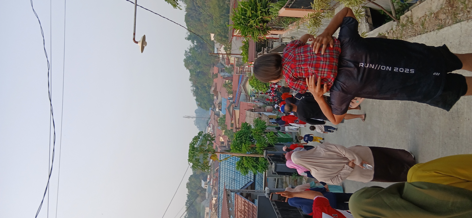 A medium shot, taken from a slightly low angle, captures a street scene in Samarinda, Indonesia. The foreground is dominated by the back of a person carrying a child on their shoulders. The child is wearing a red and black checkered shirt. The adult is wearing a black t-shirt with white text that reads "RUN//ON 2025". The street is paved and slopes downwards, with a crowd of people walking away from the viewer and down the street. Many of the people are wearing casual clothing, and some women are wearing headscarves.
The midground reveals a densely populated residential area with houses featuring red tile roofs and some blue roofs. Trees and vegetation are interspersed among the buildings. In the distance, rolling hills covered in trees form the backdrop to the scene. Overhead, power lines are visible against a hazy, light-colored sky, indicative of a possibly warm or humid day. A street lamp on a pole is also visible on the right side of the image. The overall impression is of a community gathering or procession moving through a typical Indonesian neighborhood.