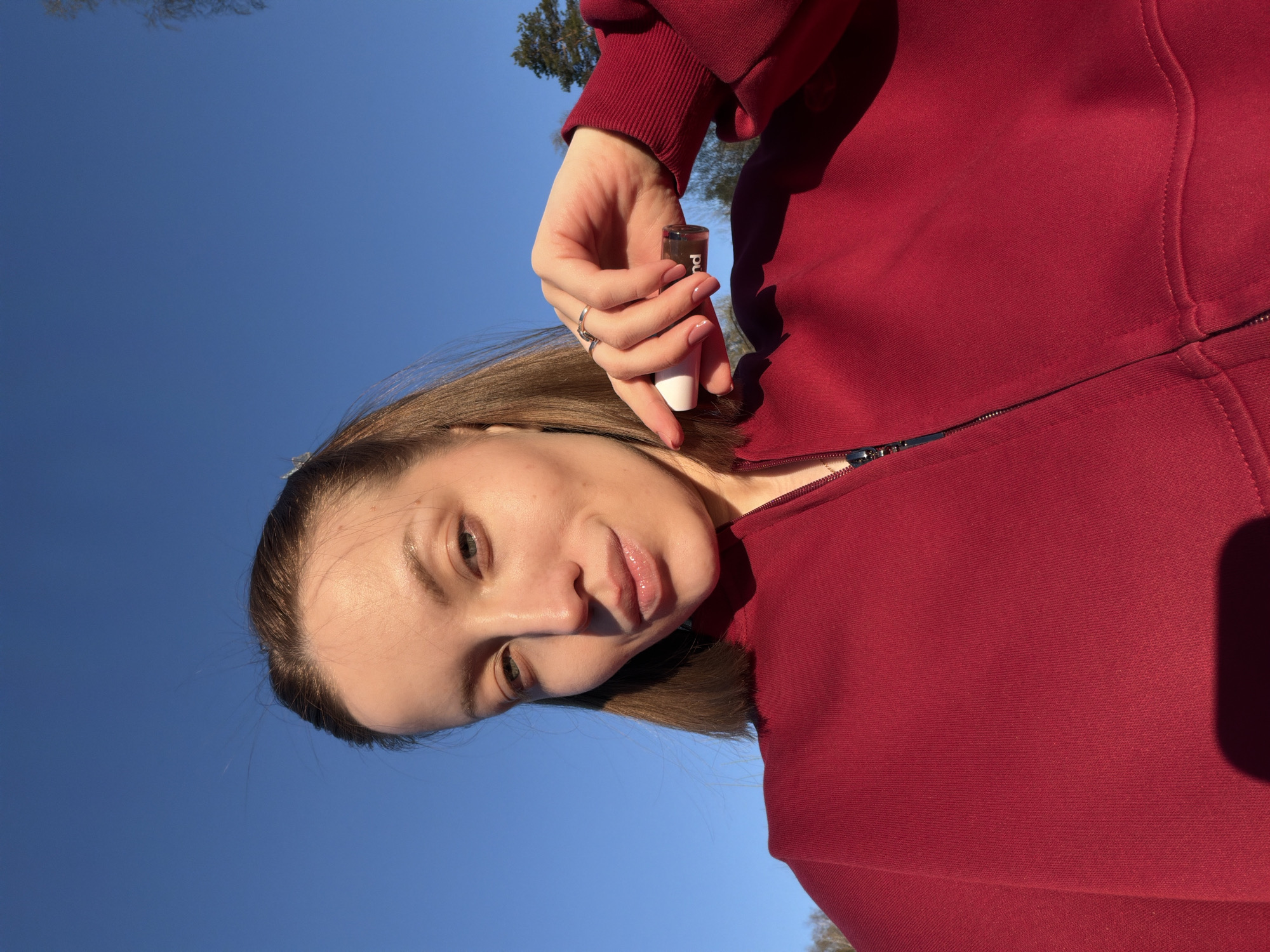 A woman is holding a small bottle of brown liquid with a white applicator wand. She is wearing a red hooded sweatshirt with a zipper. The background is a blue sky with some trees visible. The location is possibly in or near Reshetnikovo, Russia, but this cannot be definitively confirmed from the image.