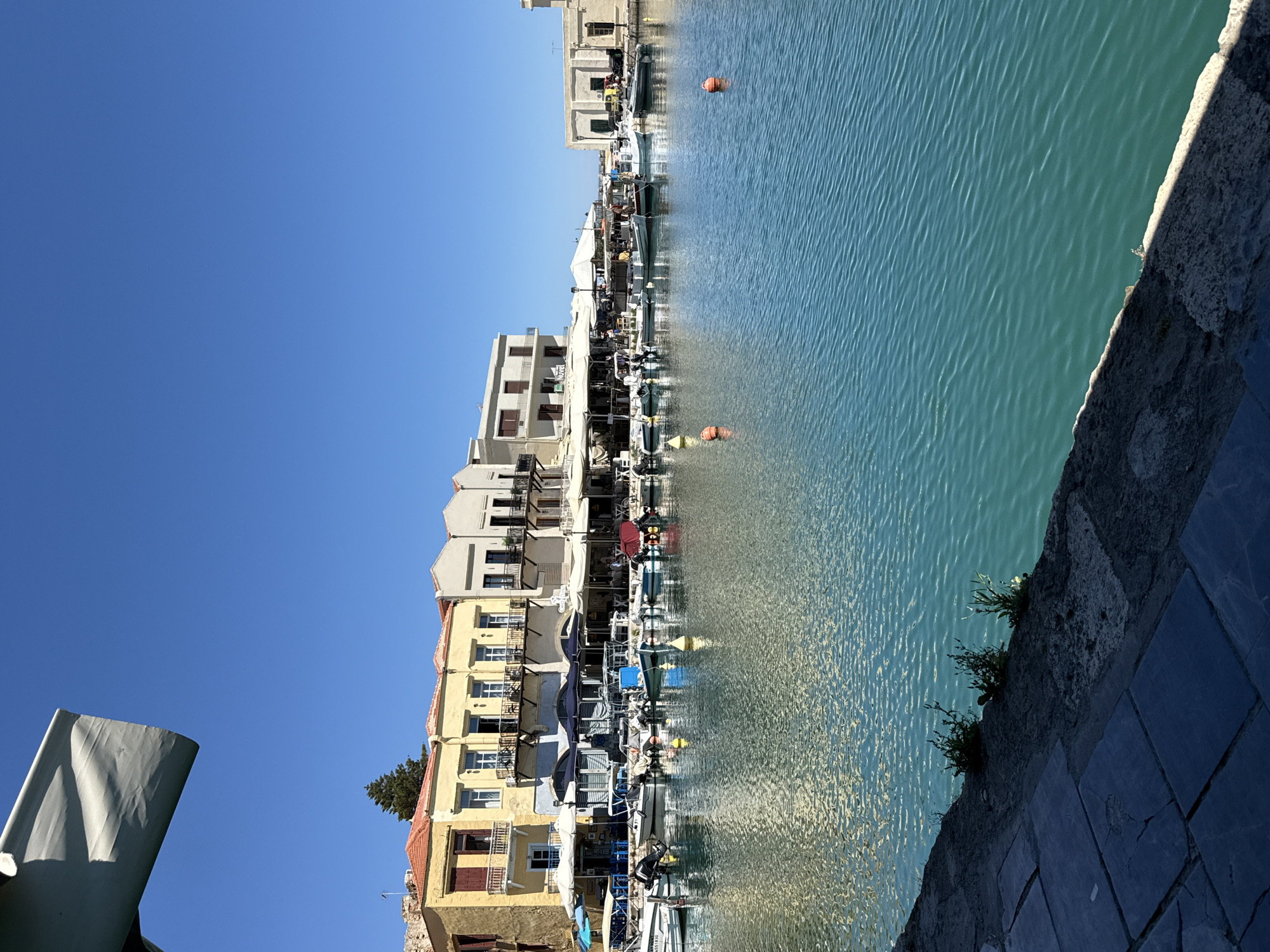 This is a vertical, eye-level, outdoor, daytime photograph of the Rethymno Venetian harbor in Rethymno, Greece. The harbor is filled with clear, turquoise water, with a ripple texture. Several small boats are docked along the waterfront. Behind the boats, a row of colorful, multi-story buildings with awnings and outdoor seating line the harbor. The buildings are light yellow, white, and beige. The sky is a clear, bright blue. In the foreground, a stone walkway and a white awning are visible. The overall scene is serene and picturesque, evoking a sense of Mediterranean charm.