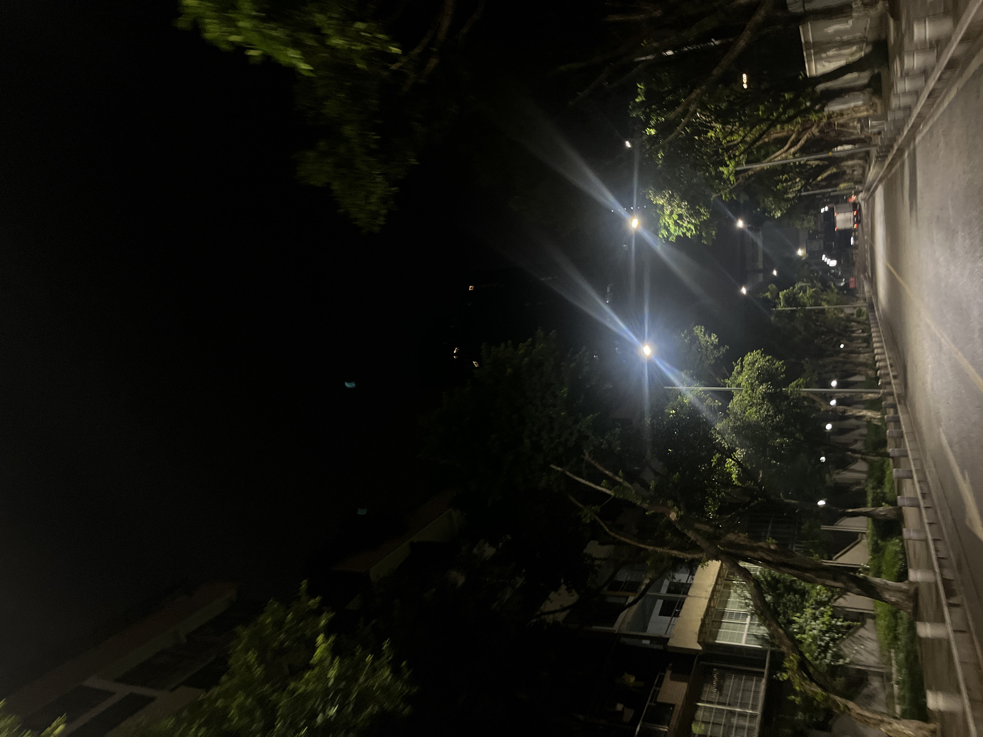 The image presents an urban street scene at night in Jiulongpo District, China. A multi-lane asphalt road, appearing somewhat reflective, recedes into the background, marked by a faint yellow median line. On both sides of the roadway, a raised concrete curb supports a series of evenly spaced cylindrical bollards. Dense foliage and visible trunks of mature trees line both edges of the street, forming a canopy that partially obscures the upper portions of the scene. Multiple overhead streetlights illuminate the area with bright white light, generating prominent lens flares. To the left, multi-story buildings, featuring balconies and windows, are partially visible behind the tree line. On the right, a boundary wall or fence runs parallel to the street, also partially hidden by trees. A white, enclosed vehicle, consistent with a truck or van, is discernible in the distant background, oriented away from the viewer. The sky above is dark, with minimal distant light sources visible from taller structures. No individuals are present in the observable environment.