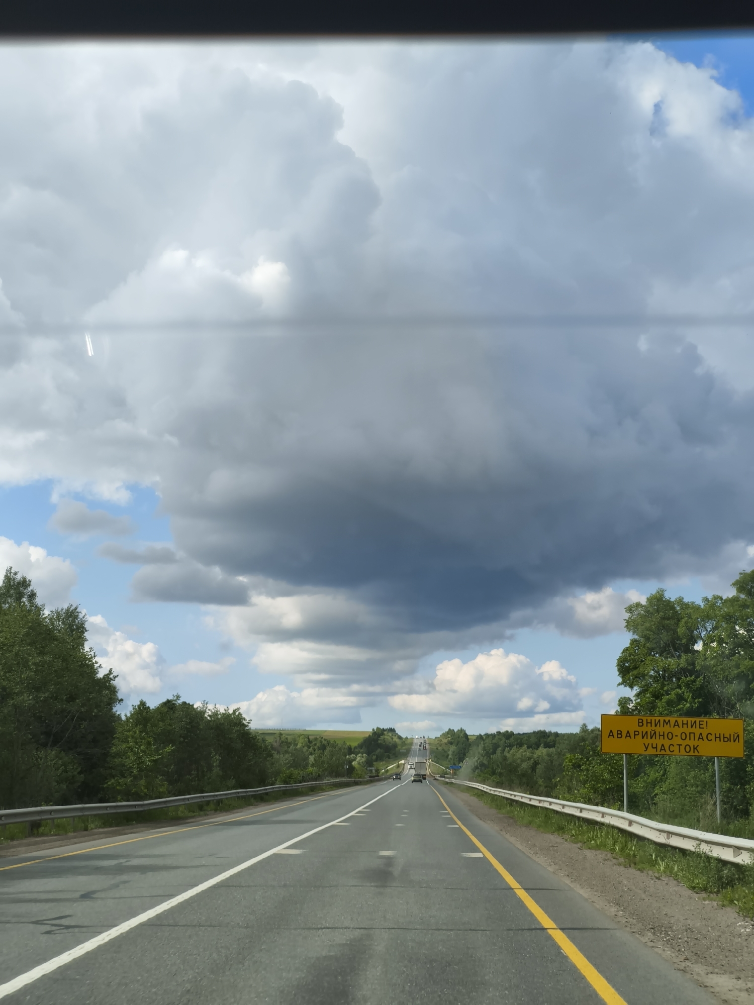 The image captures a first-person perspective from inside a vehicle traveling along a multi-lane highway near Старое Мартьяново, Russia, on a day with dynamic weather. The asphalt road stretches into the distance, with clear white lane markings and a solid yellow line separating the driving direction from the shoulder on the right. A metal guardrail runs along the right side of the road, bordering a dense line of green trees and lush vegetation. More trees and fields are visible in the far distance, indicating a rural or semi-rural setting.
The sky is a prominent feature, displaying a dramatic contrast between large, dark grey storm clouds dominating the upper and central parts of the frame, and patches of bright blue sky with fluffy white cumulus clouds visible below and to the left of the main cloud mass. This suggests changing weather conditions, possibly before or after a rain shower.
On the right side of the road, a bright yellow rectangular sign is clearly visible. The text on the sign, written in Cyrillic, reads:
"ВНИМАНИЕ!" (ATTENTION!)
"АВАРИЙНО-ОПАСНЫЙ" (ACCIDENT-PRONE / HAZARDOUS)
"УЧАСТОК" (SECTION)
This sign warns drivers of a dangerous stretch of road ahead.
In the far distance, other vehicles can be seen traveling on the highway. The time of day appears to be daytime, likely late morning or early afternoon, given the natural light although the heavy clouds dim some areas. The scene depicts a journey through a landscape experiencing varied weather.