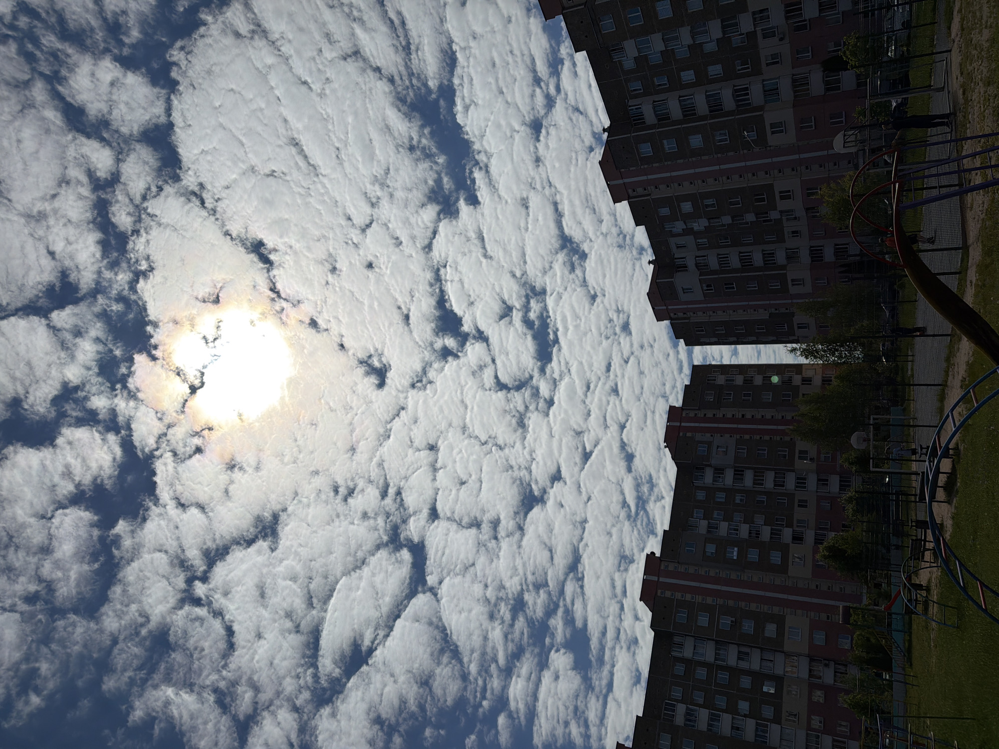 The image captures a residential scene featuring a partly cloudy sky, tall apartment buildings, and a playground area. The upper left portion of the image is dominated by dense, textured white clouds against a blue sky, with a bright, intense sun visible through a break in the clouds. To the right and bottom right, several multi-story apartment buildings rise, characterized by numerous rectangular windows and a facade primarily of brick or a similar material, with some sections painted in lighter tones. In the foreground, a grassy patch hosts playground equipment, including a red and yellow slide, a blue and red climbing structure, and a basketball hoop. Two small, dark, silhouetted figures, likely people, are visible near the basketball hoop. Trees with green foliage are scattered between the buildings and near the playground. The visible text includes a large number "7" painted on a lighter section of one of the buildings. The time of day appears to be daytime, with bright sunlight and clouds creating varied illumination.