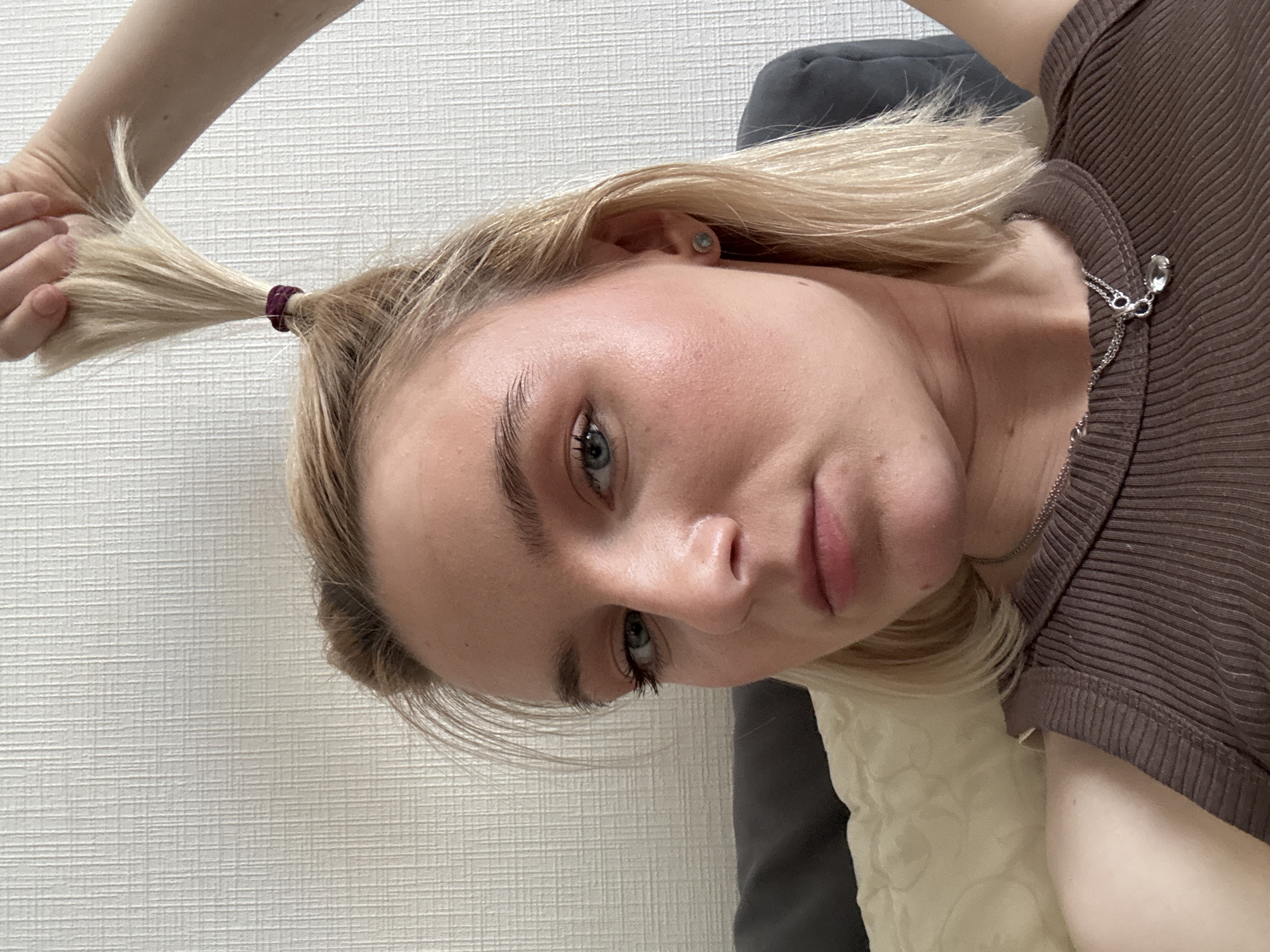A young, fair-skinned woman with light blonde hair is looking into the camera with a slight tilt of her head. Part of her hair is gathered in a small ponytail, held with a dark-colored hair tie. Another hand is holding the ponytail further up. She has blue eyes, light eyebrows, and pink lips. She is wearing a brown ribbed top. A silver necklace is visible around her neck. A white wall with a subtle textured pattern is in the background. Part of a gray cushion or pillow is also visible. The photo does not appear to be taken in Vsevolozhsk, Russia, as there are no elements that suggest this location.