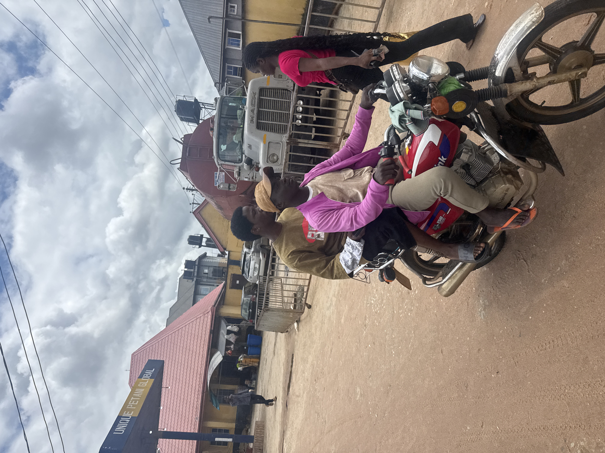 A dusty, unpaved road in Ihiagwa, Nigeria, is the setting for this image. Three young adults are prominently featured. Two are on a red motorcycle. The person driving wears a light brown t-shirt, tan pants, and a bright pink jacket. They are wearing orange flip-flops. The passenger behind them wears a brown long-sleeved shirt. A young woman with long braided hair, wearing a pink t-shirt and black pants, stands to the right of the motorcycle, facing the driver. In the background, there is a red tanker truck with "MACK" written on its front. A building with a pink roof, bearing the sign "UNIQUE PETANI GLOBAL," is visible on the left. Other buildings, a metal fence, and a few parked vehicles are also present. The sky is overcast with dramatic clouds.
