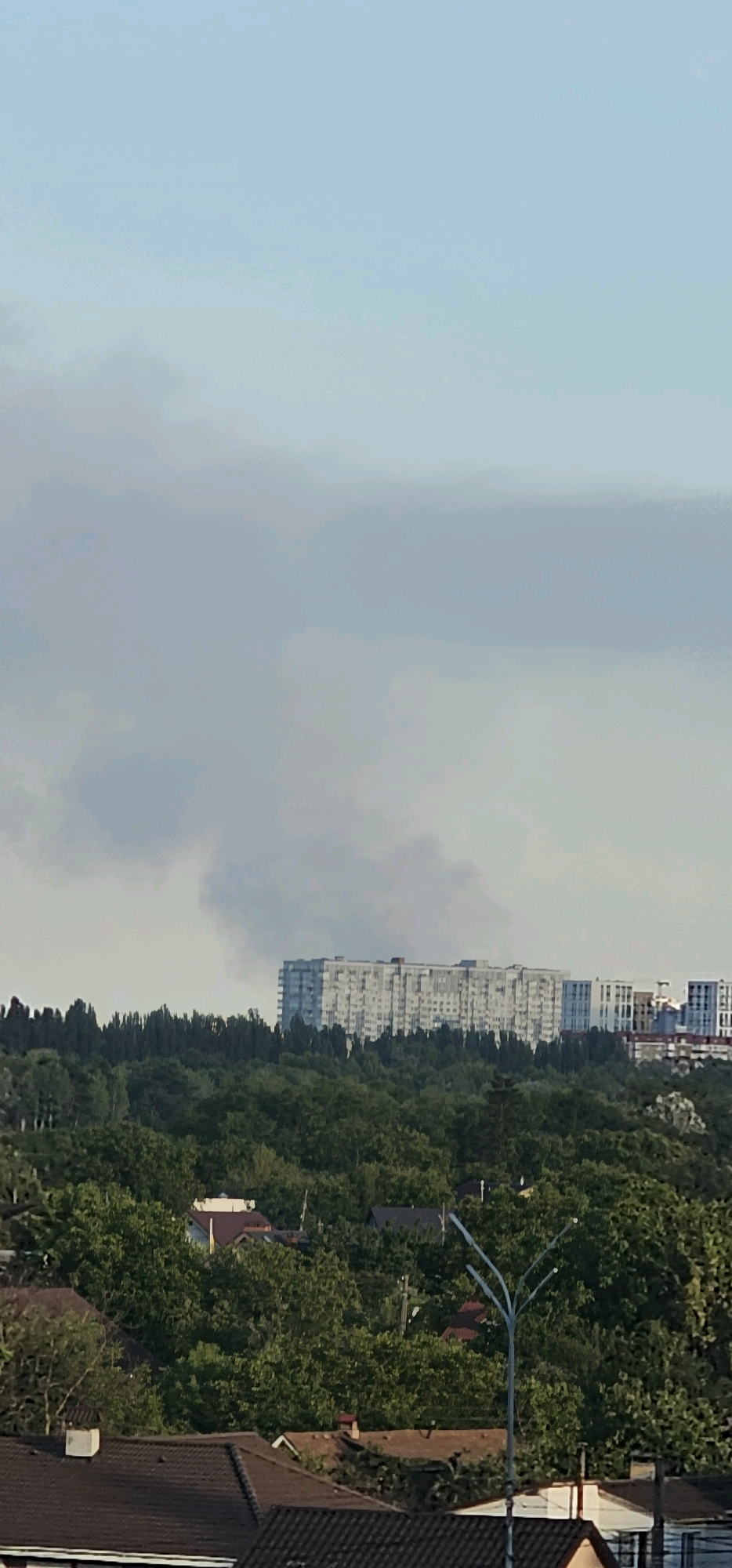 A vertical, medium-shot, outdoor photograph shows smoke rising from behind a dense forest and a row of houses in the foreground. In the midground, a long, multi-story building, possibly an apartment complex, is visible. Behind it, a plume of dark smoke is rising into the sky. The sky is overcast with light clouds. The environment is urban with elements of nature. The image is not in focus.