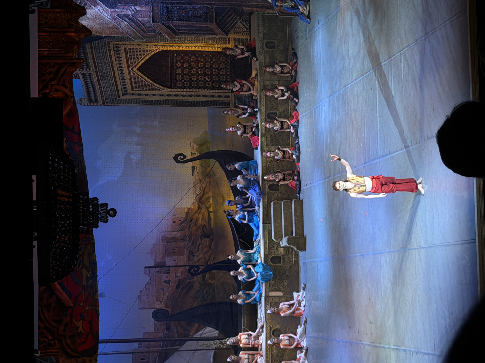 The image captures a theatrical performance, possibly a ballet or opera, taking place on a stage. A backdrop depicts a city scene with architectural details, including arches and sail-like structures. Onstage, performers in various costumes are arranged; some wear blue garments and sit, while others wear red and gold outfits and appear to be lying or dancing. The primary dancer is positioned in the foreground, reclined on the stage with their arms outstretched. The stage floor is a light blue color with panel lines. The location is the State Opera and Ballet Theatre of Udmurtia in Izhevsk, Russia.