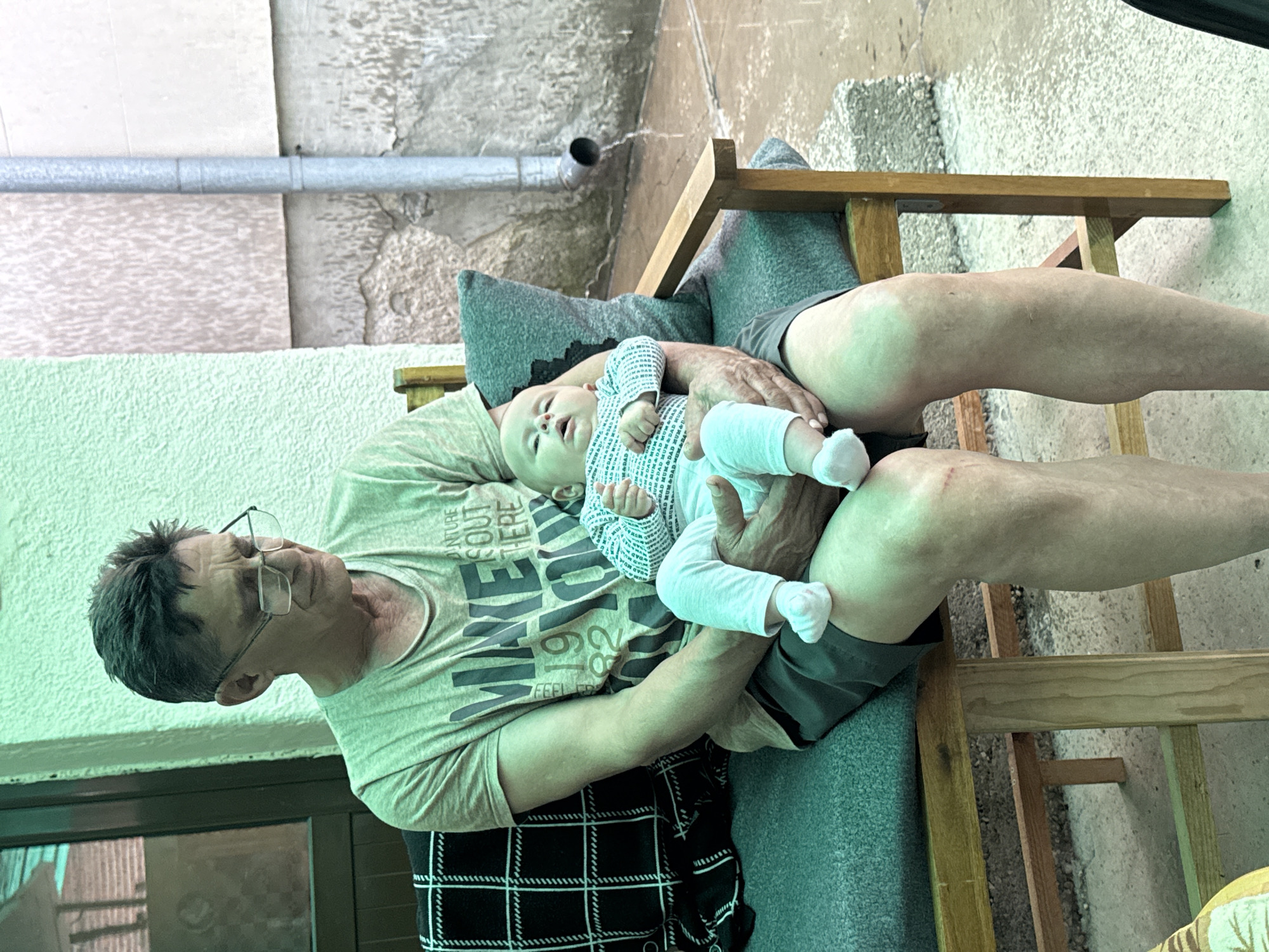 An older man wearing glasses, a light-colored t-shirt, and shorts sits on a wooden chair with grey cushions, holding an infant on his lap. The man is looking down at the baby with a gentle expression. The baby, dressed in a white and blue patterned onesie and light-colored socks, looks upwards. The man's hands gently support the baby's head and body.
The setting appears to be an informal, possibly covered outdoor area or an interior room with rustic elements. The background features textured walls, possibly concrete or stone, with varying shades of grey and beige. A horizontal metal pipe runs across the upper part of the wall. To the left, a dark window or door frame is visible. A plaid black and white blanket or fabric is draped over the man's lap and the chair.
Visible text on the man's t-shirt includes fragments such as "NTURE," "S OUT HERE," "EL YOU," and "FEEL FR," part of a larger slogan. The baby's onesie has a repeating, small-scale pattern, too indistinct to read clearly. The lighting is soft and diffused, suggesting daytime in a shaded area. The scene conveys a quiet moment of bonding between the man and the infant. The overall atmosphere is relaxed and intimate.