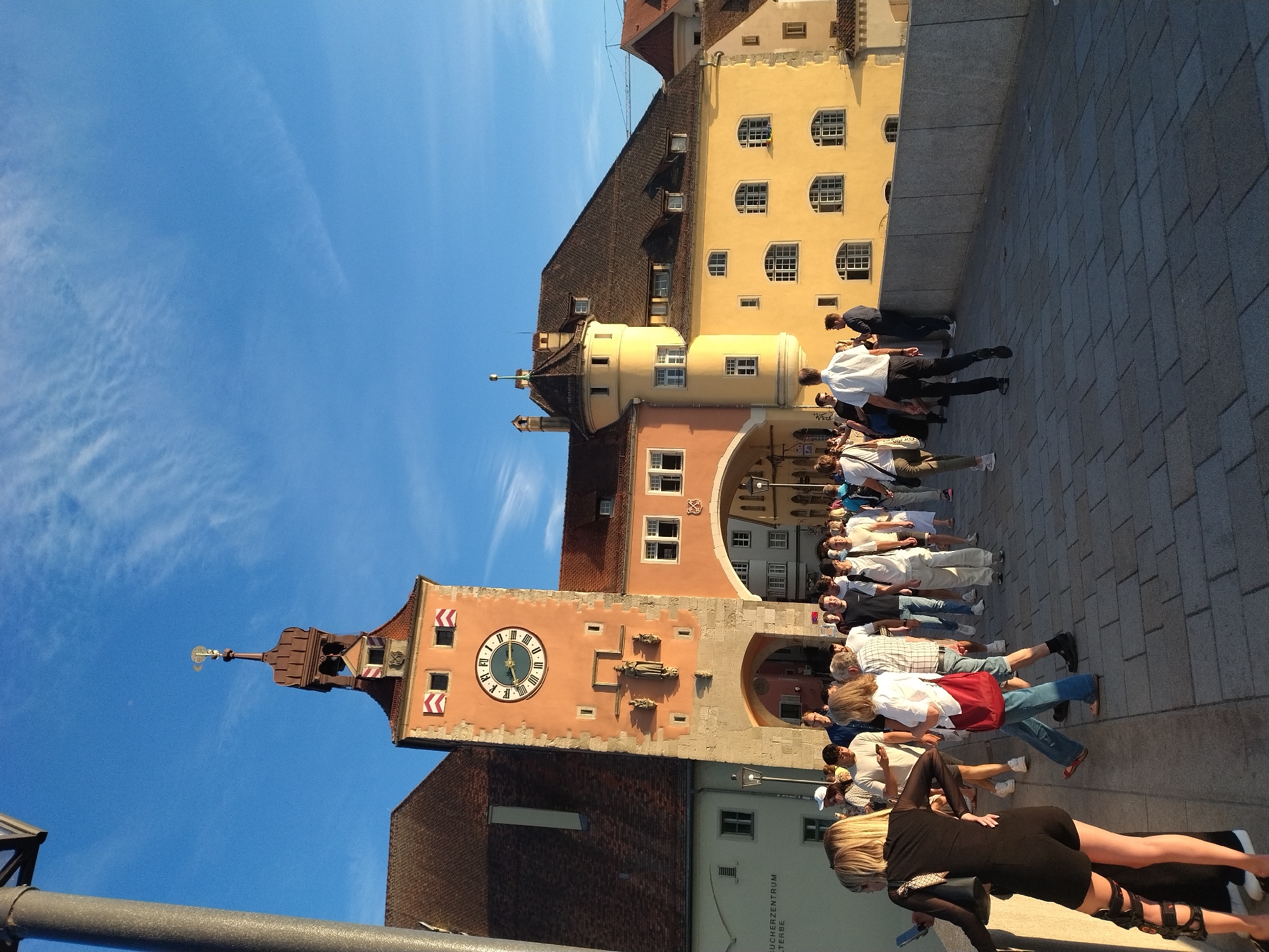 A street scene unfolds in Regensburg, Germany, likely during the late afternoon given the warm, low-angle sunlight casting long shadows. In the background, a prominent, peach-colored tower with a clock face dominates the view. The tower features Roman numerals on its clock and is topped with a dark, multi-tiered roof and a golden weather vane. Adjacent to the tower is a bright yellow building with arched windows and a dark tiled roof, and to the left, a darker building with a steeply pitched roof. A stone bridge with a grey railing spans across the foreground, where a crowd of pedestrians is walking.
The people are dressed in casual summer attire, suggesting a pleasant temperature. They are moving in various directions, some in small groups, others individually. One woman in the immediate foreground on the left is wearing a black dress and poses, looking towards the left. Another woman to her right, wearing a white shirt and blue jeans, carries a red bag. Several men are visible, some in t-shirts and jeans, others in button-down shirts. The pedestrian bridge is paved with large, rectangular grey stone tiles. The sky above is a clear, vibrant blue with wisps of white clouds. The architecture and the bridge suggest a historical district within the city.