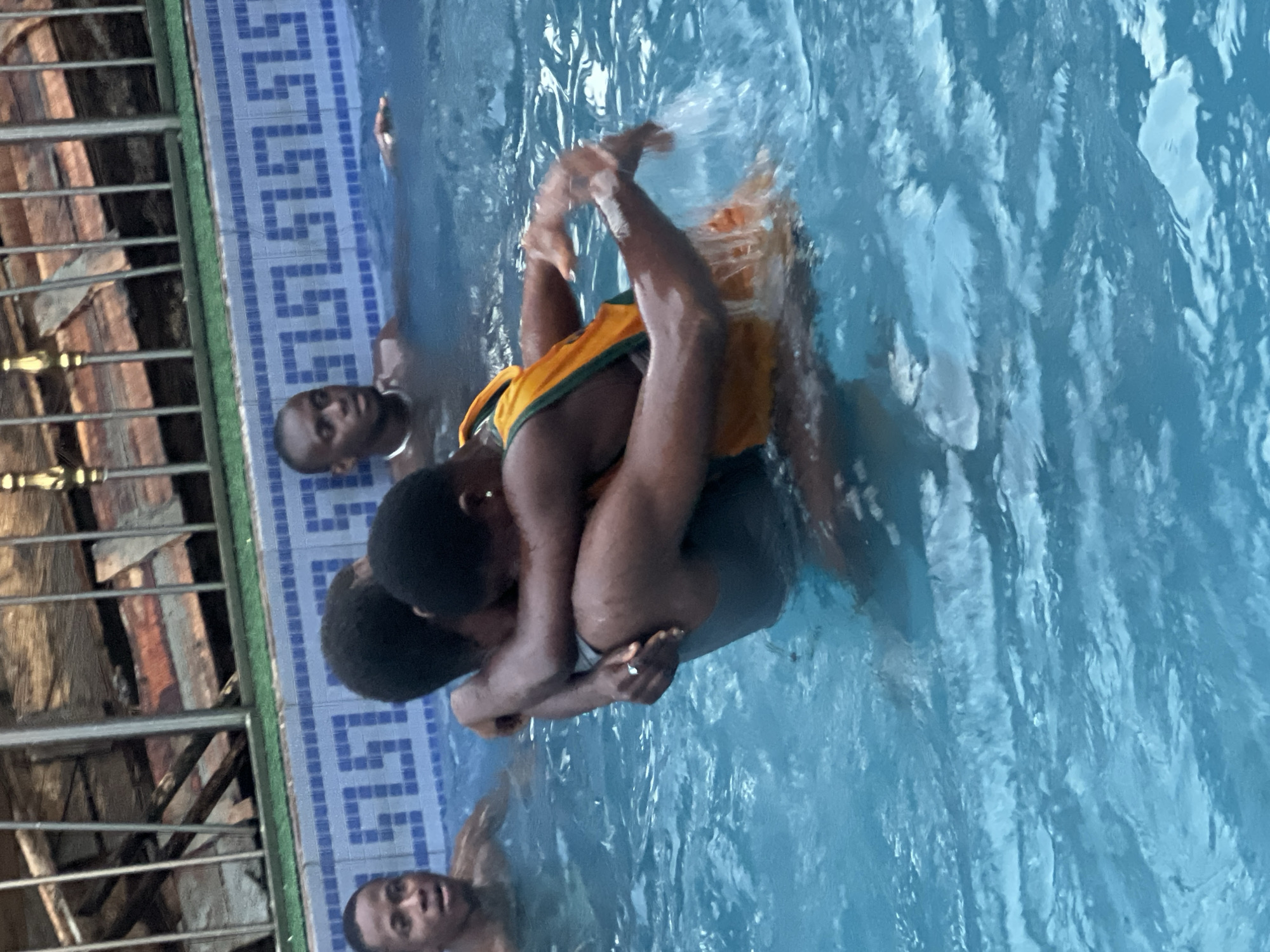The image depicts a lively scene in a swimming pool in Nkpor, Nigeria. Several dark-skinned individuals are present in the blue water. At the center, two individuals are closely interacting; one is holding the other, who is wearing a yellow and green top, suggesting a playful embrace or interaction. Other individuals are partially visible in the water in the background and foreground. The water is agitated, indicating active movement and splashing. The pool's edge is adorned with distinctive blue and white patterned tiles. Outside the pool area, a metal fence and parts of a building with visible brick or concrete are discernible. The lighting suggests it is daytime. No visible text is present in the image.