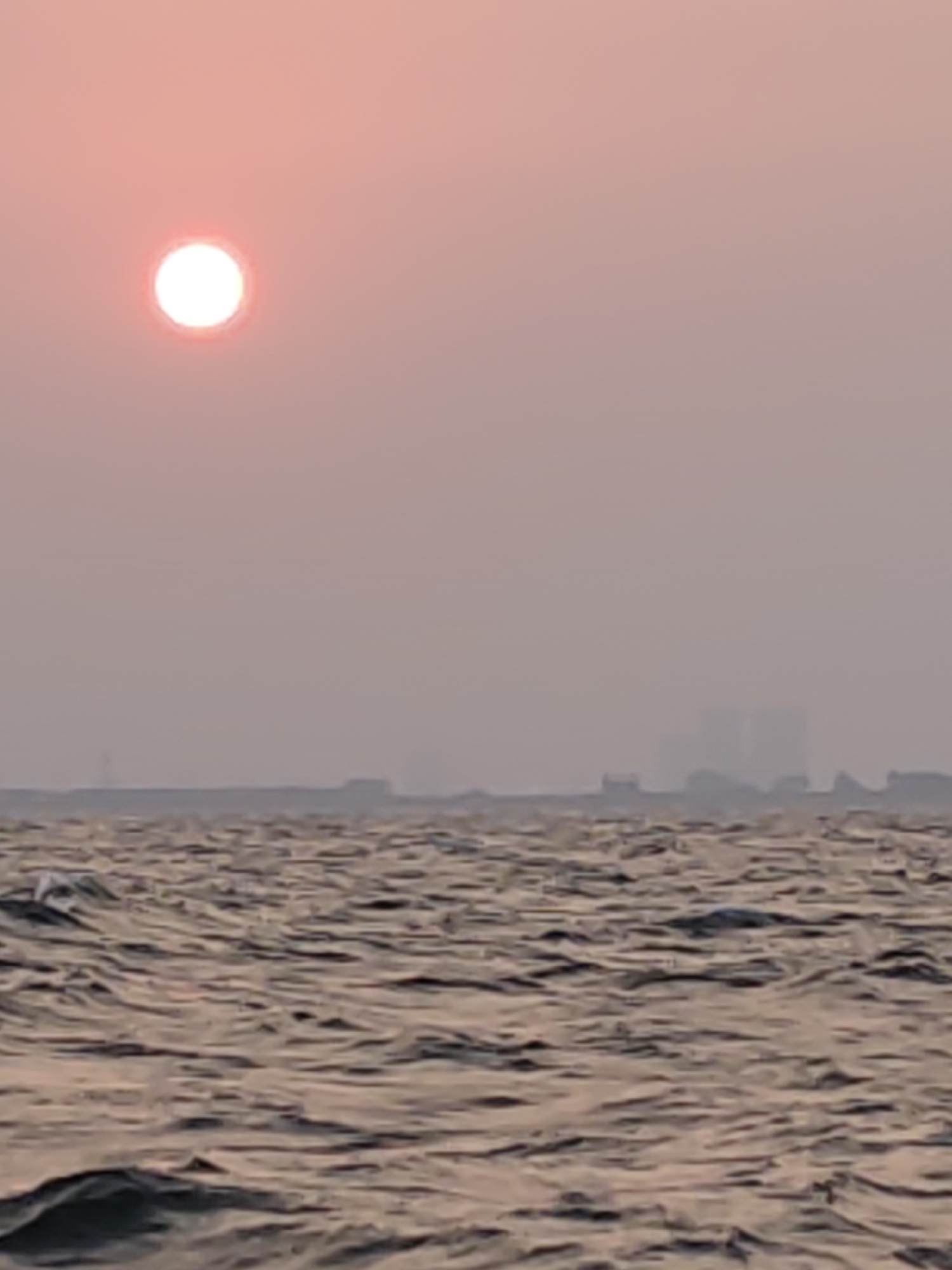 A wide shot captures a hazy sunrise over a body of water. A bright, glowing orb, presumably the sun, is positioned in the upper left quadrant, casting a soft, pinkish hue across the sky. In the distance, a blurred, silhouetted city skyline is visible, with indistinct buildings emerging from the mist. The foreground is dominated by choppy water, reflecting the muted light of the sunrise with ripples and waves creating a textured surface. The overall scene is atmospheric and serene, with a sense of quietude.