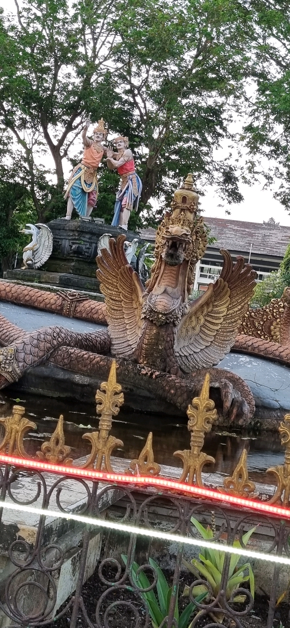 The image depicts a statue of a mythical creature with bird-like wings and a dragon-like head, possibly a Garuda or Naga, in the foreground. The statue is ornate, with intricate detailing on its scales, wings, and headdress. It appears to be part of a larger decorative fountain or sculpture. In the background, there are two humanoid statues dressed in traditional Balinese attire, one with an orange top and blue skirt, the other with a red top and blue skirt. They stand on a tiered stone platform, possibly depicting a scene from Balinese mythology. The statues are set against a backdrop of lush green trees and a building with a tiled roof. The scene is illuminated by decorative LED lights running along a fence in the foreground, casting a red and white glow. The overall impression is of an outdoor cultural or religious monument, likely located in Denpasar, Indonesia, given the Balinese architectural and sculptural style.