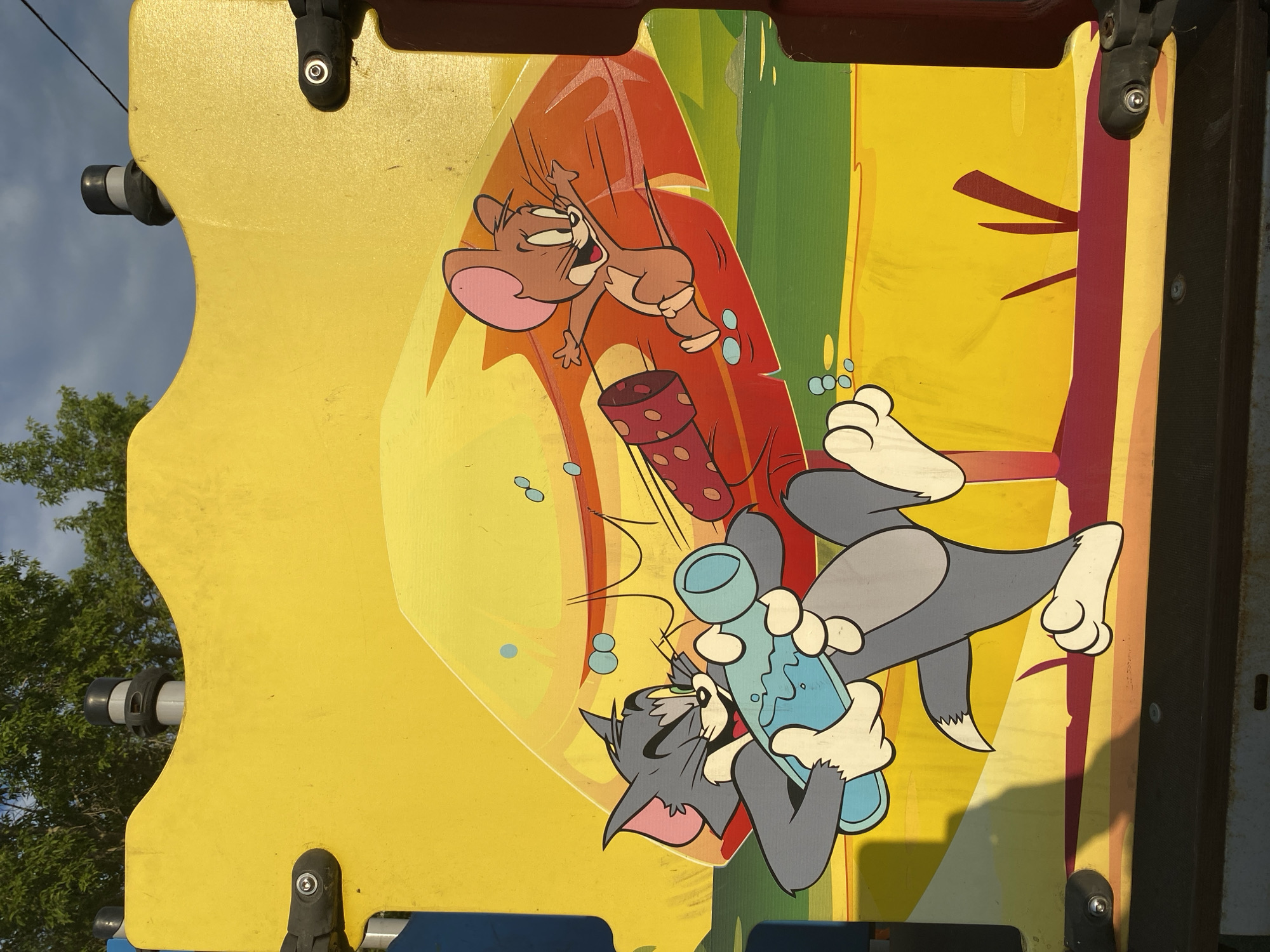The image shows a colorful mural featuring cartoon characters Tom and Jerry on a yellow panel, likely part of a playground structure. Tom, a gray cat, is depicted holding a blue bottle with liquid, and Jerry, a brown mouse, is shown running away from a red, polka-dotted object. The mural is set against a backdrop of green and yellow stripes, and the sky is visible above with trees in the upper left corner. The setting appears to be outdoors, with sunlight casting shadows on the mural. The mural is located at a playground in Petropavl City Administration, Kazakhstan.