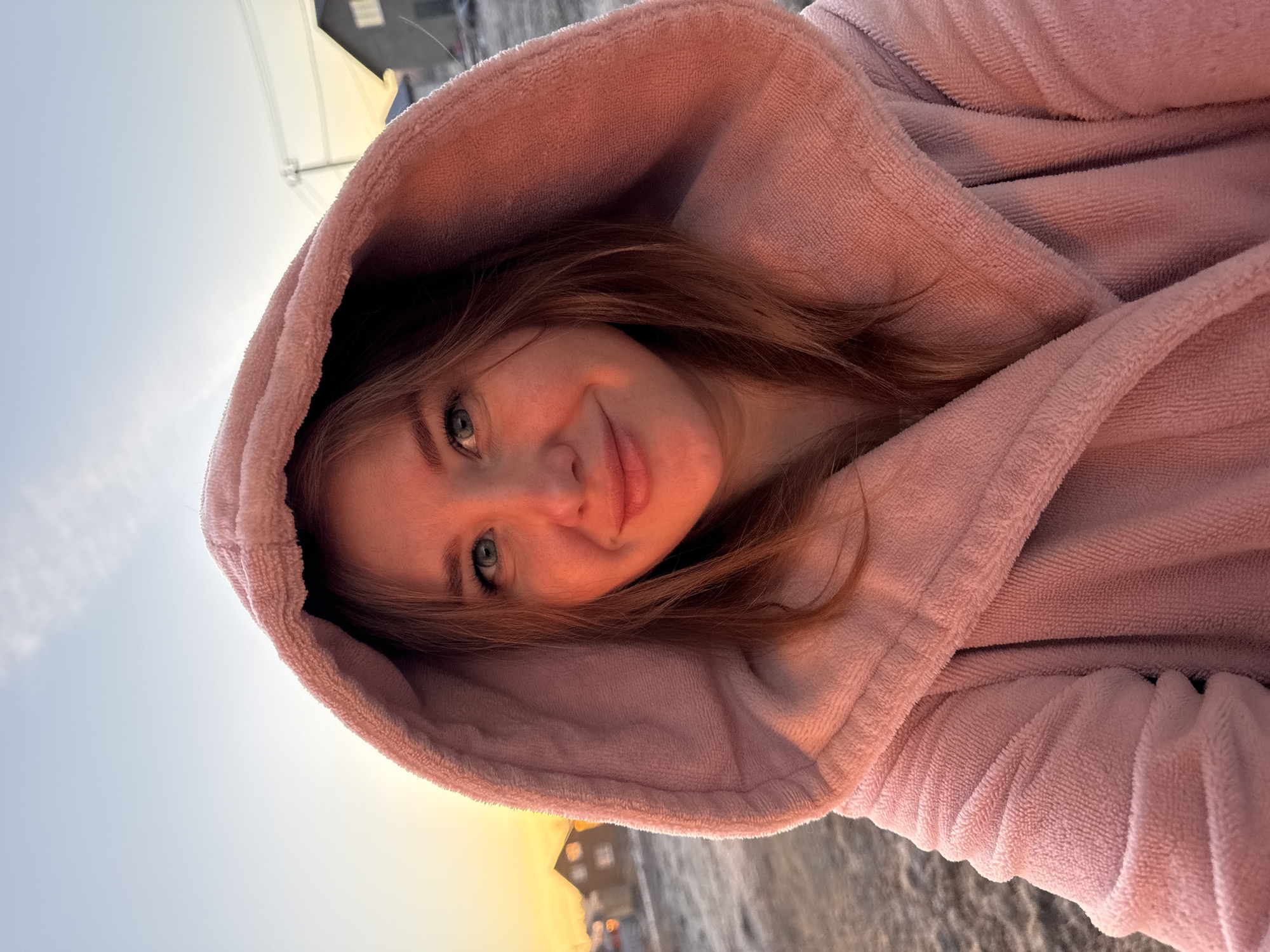 A young woman with long, light brown hair is wearing a light pink hooded robe. She is smiling slightly and looking directly at the camera. The background is slightly blurry, but appears to be an outdoor setting with a building in the distance. The overall image has a soft, warm tone, possibly taken during sunrise or sunset. Gornyi (Dvurechenskoe S/P), Russia.