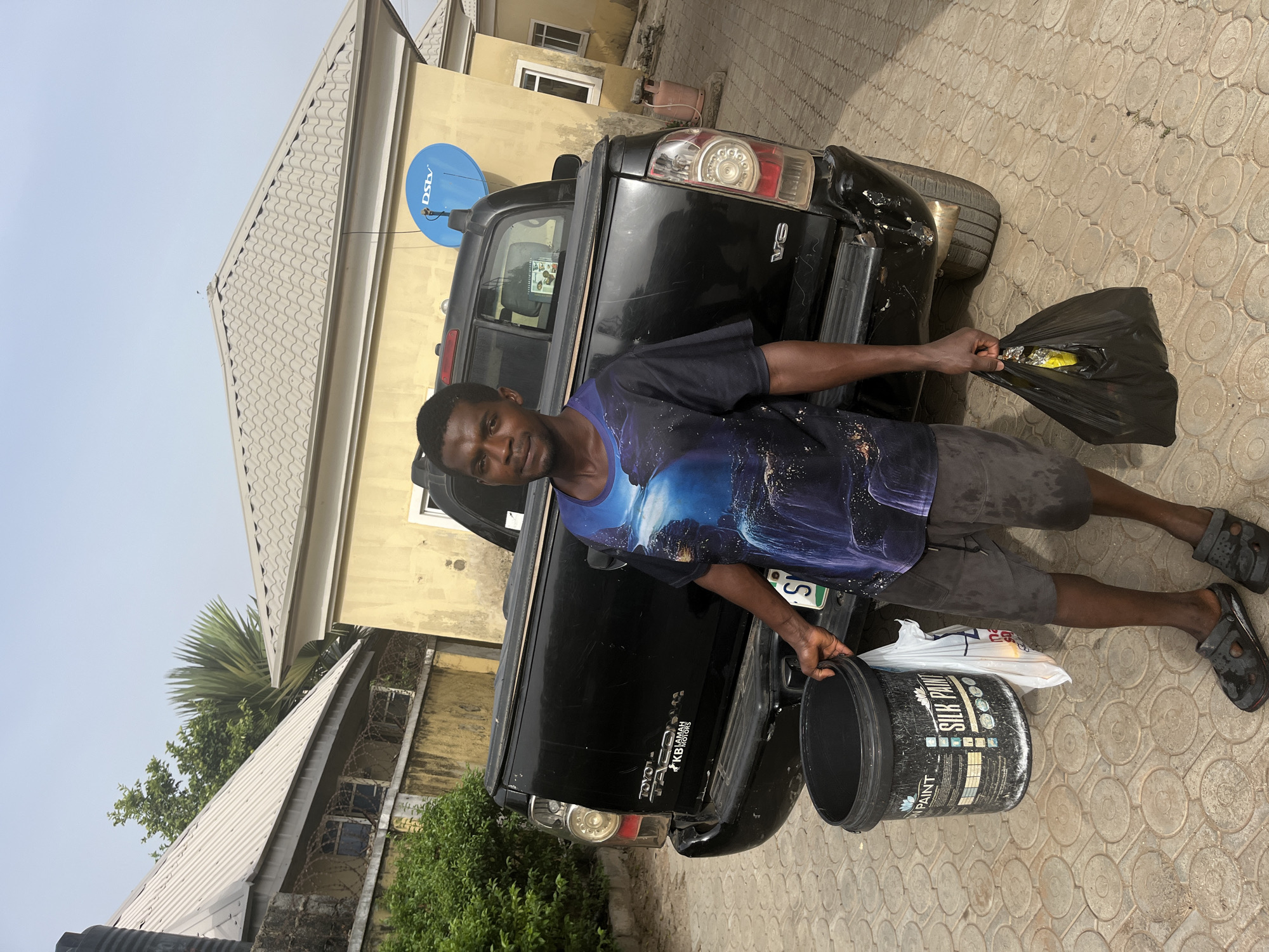 A man stands next to a black pickup truck. He is holding a bucket of "Silk Paint" in his left hand and a black plastic bag in his right hand. The truck is parked on a paved surface with a repeating circular pattern. Buildings are visible in the background, including a yellow house with a blue satellite dish. Green foliage is also present. There is no explicit information indicating that the location is specifically Abuja, Nigeria, but the license plate on the truck is Nigerian.