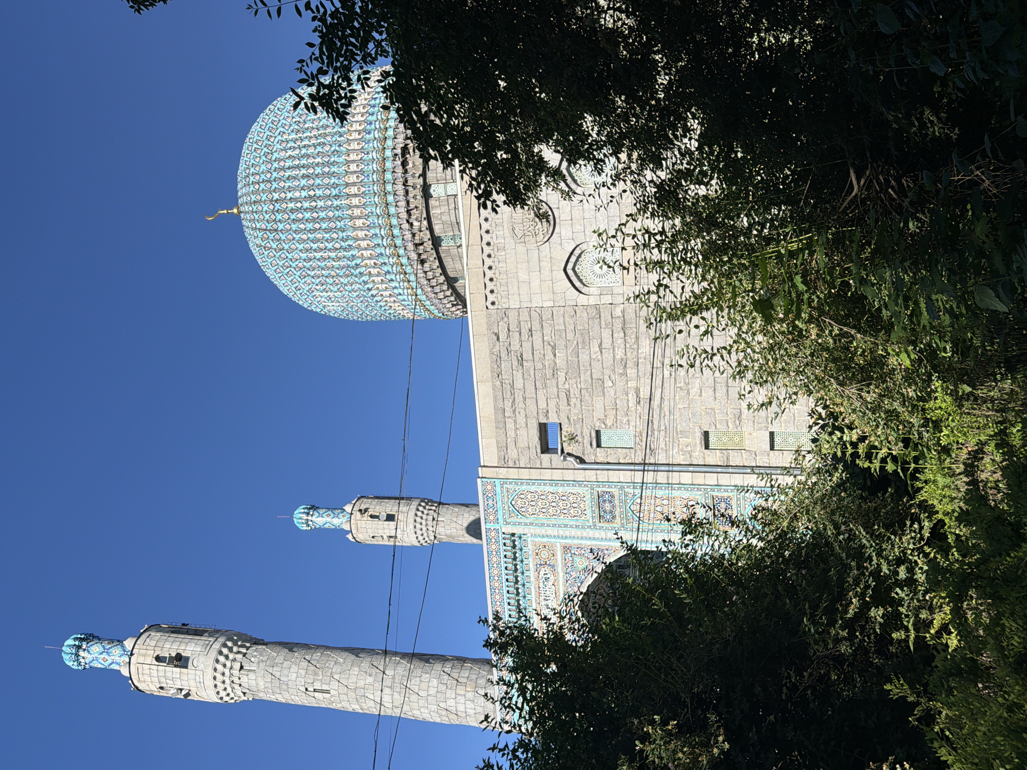 This image depicts the Saint Petersburg Mosque, located in Saint Petersburg, Russia, specifically at Kronverksky Prospekt, 7. The structure is observed from a low angle, with dense green foliage partially obscuring its lower portions.
The mosque features a large, spherical main dome prominently covered in intricate mosaic tilework, primarily in shades of blue, white, and gold, arranged in geometric patterns. A golden crescent moon finial surmounts the dome. Two cylindrical minarets are visible; the taller one is positioned on the left, and a shorter one is partially visible behind the main building section to the right. Both minarets are constructed of light grey, textured stone blocks and feature decorative blue and white tiled sections near their apexes, mirroring the dome's patterns. Each minaret includes small, vertically oriented windows.
The main building facade is primarily composed of light grey, rough-hewn stone blocks. A significant section of the facade, particularly on the left side, is adorned with elaborate multi-colored tilework featuring geometric and calligraphic designs in hues of blue, white, green, and gold. Other stone sections of the facade exhibit arched windows with white decorative grilles and, on the right side below the dome, a circular stone carving containing Arabic script. Thin, dark horizontal lines, likely utility cables, traverse the mid-ground of the image.
The sky above is clear and bright blue, occupying the upper third of the frame. Sunlight illuminates the mosque and parts of the foliage, creating distinct areas of shadow within the dense greenery in the foreground and mid-ground. No individuals are visible, nor are any specific actions depicted.