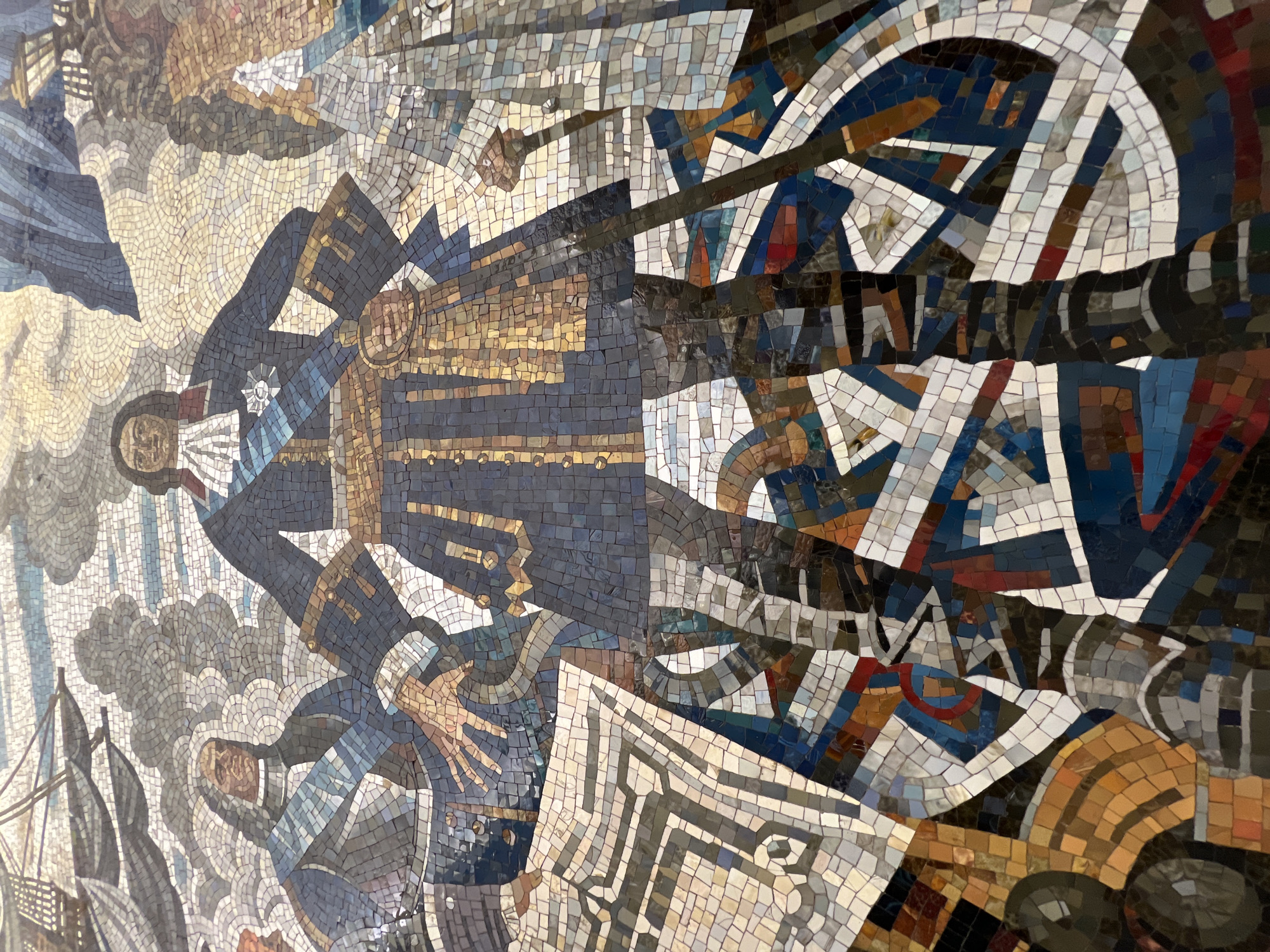 This is a mosaic depicting a naval battle scene. The mosaic is located in the Saint Petersburg Metro, Russia.
The scene shows military figures in naval uniforms, likely officers, in a dynamic pose. There are ships and cannons depicted amidst smoke and explosions. The mosaic utilizes a variety of colored tiles to create a textured and detailed representation of the battle.