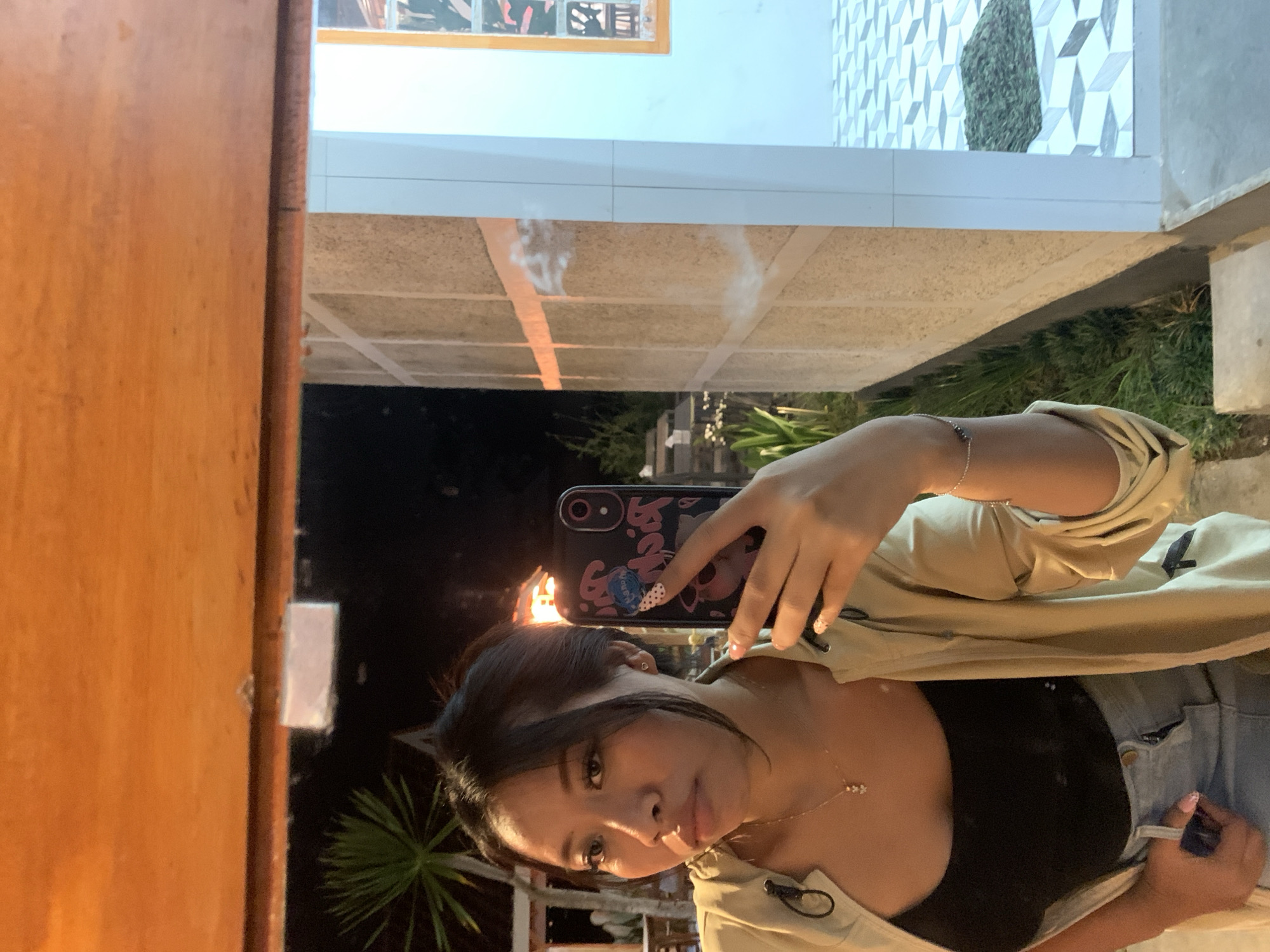 In Jajag, Indonesia, a young woman is captured in a mirror selfie at night. She is wearing a black tank top under an open tan jacket and blue denim shorts. A delicate necklace with a small charm adorns her neck. She holds a patterned phone in her right hand, taking the picture. Her left hand rests on her pocket. The background reveals an outdoor setting with a textured wall and tiled flooring, suggesting a pathway or patio area. A palm tree and some foliage are visible in the distance, along with a hint of building structures and lighting that indicates a residential or commercial area. The overall ambiance suggests a casual evening setting.