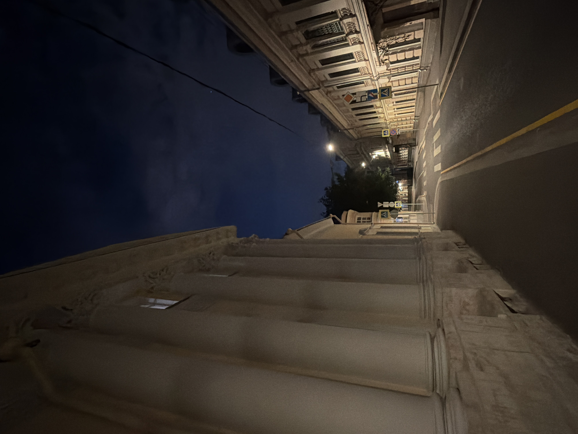 The image depicts a street scene at night in Moscow, Russia, specifically on Chistye Prudy Boulevard. The left side of the frame is dominated by a large, light-colored building with prominent, classical-style columns. The right side of the street features a row of cream-colored buildings with dark-framed windows and decorative architectural details. The street itself is paved, with visible white pedestrian crossings and yellow lane markings. Several yellow traffic signs with blue symbols are present along the sidewalk, indicating pedestrian crossings. Streetlights cast a warm glow on the scene, illuminating the road and the facades of the buildings. The sky is dark blue with scattered clouds, and a thin power line stretches diagonally across the upper portion of the image. No people are visible in the frame.