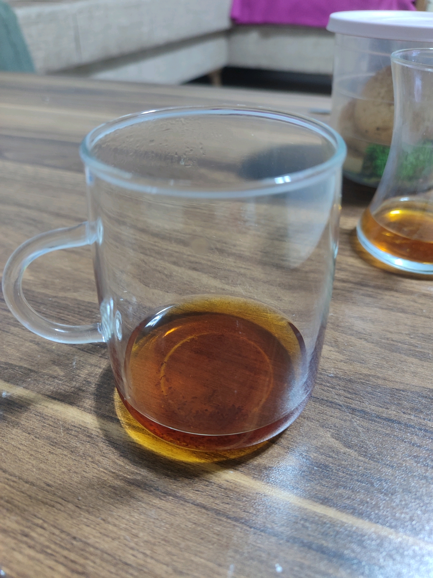A clear glass mug with a handle contains a small amount of amber liquid, likely tea. The mug is positioned on a wooden table with a visible grain. In the background, out of focus, are two clear containers. One appears to be a cylindrical jar with a white lid, and the other is a glass flask holding a similar amber liquid. The overall scene suggests a casual indoor setting.
