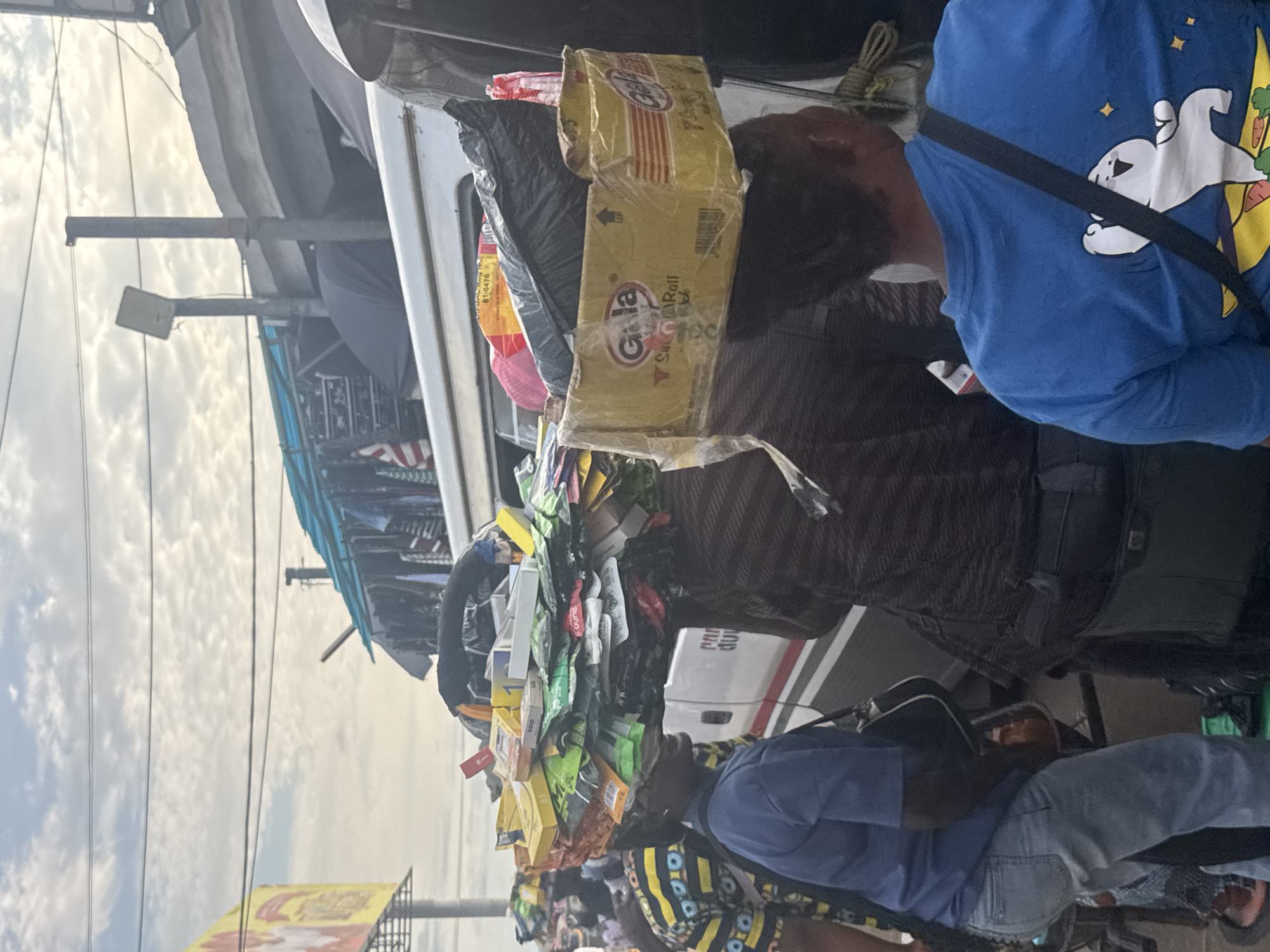 In Onitsha, Nigeria, an individual is captured with a substantial assortment of goods balanced precariously atop their head, including a yellow cardboard box labeled "Gala Salt'n'Roll" and numerous smaller packaged items. The person is wearing a blue t-shirt with a cartoon character and a black crossbody strap, and light-wash jeans. They are standing next to a white vehicle with red and black lettering that reads "pany dulj". Another person, wearing a patterned shirt and a blue top, stands to the left, partially obscuring the view of the vehicle. In the background, a sky filled with scattered clouds suggests it is daytime. Overhead, power lines crisscross the sky, and in the distance, a structure covered with a blue tarp, likely a makeshift stall or shelter, is visible, with various items, possibly clothing, hung or displayed. The scene depicts a busy street or market environment, characteristic of commercial activity in the city.