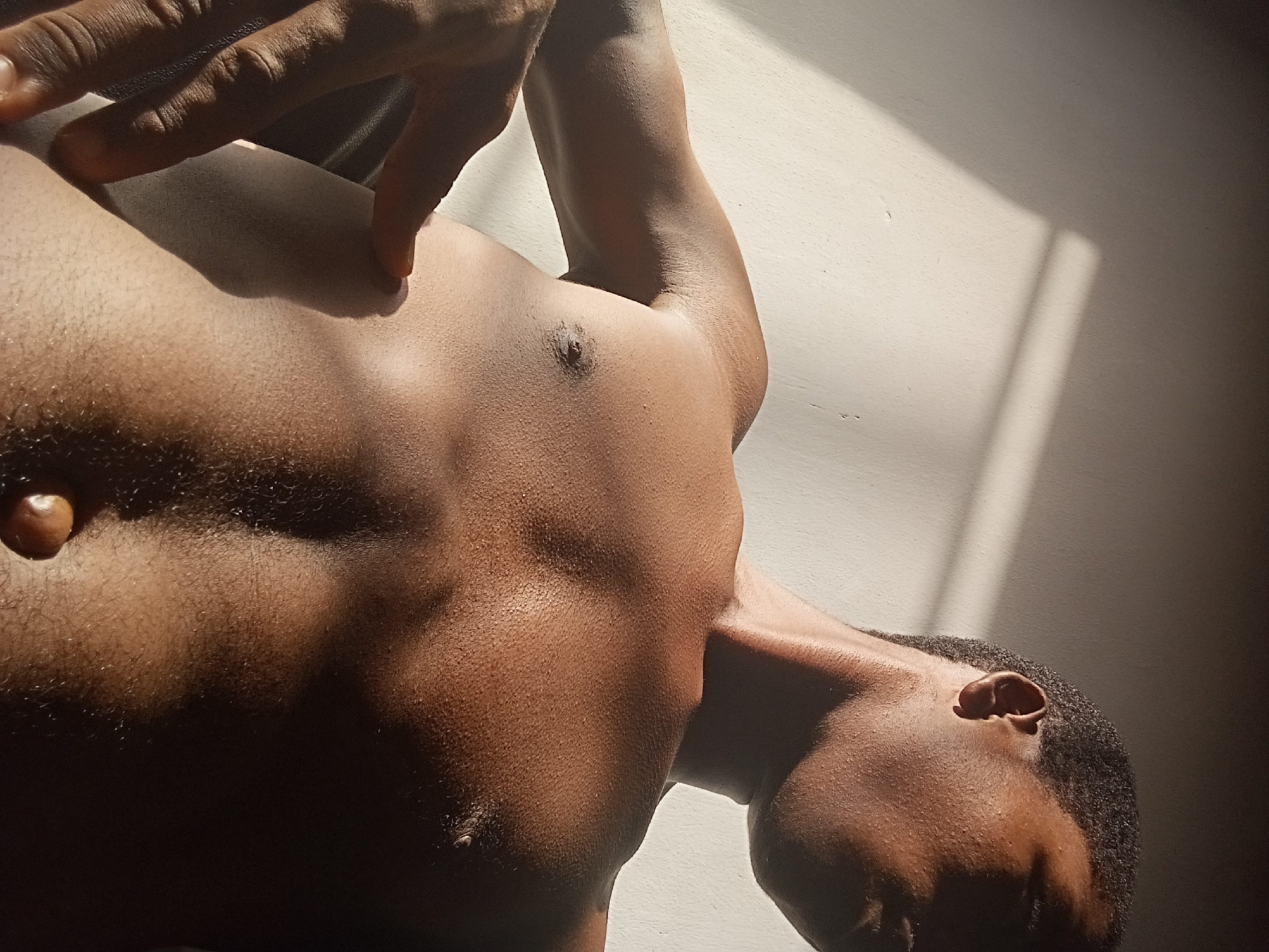 A close-up, high-angle shot captures the upper body of a shirtless person with dark skin, positioned against a light-colored background. The individual's body is angled, with their head partially visible in the lower right and their torso extending diagonally upwards to the left. Their right arm is raised, with the hand resting on or near their hip, casting a distinct shadow.
Strong, directional natural light, likely sunlight, illuminates portions of the person's chest, shoulder, and arm, creating bright highlights and deep, contrasting shadows. The light emphasizes the contours of their muscular physique. Visible details include short, dark, curly hair around the ear and head, and fine, dark hair on the chest and abdomen. A noticeable umbilical hernia is present on the abdomen.
The background appears to be a plain, light-colored wall or surface. Sharp, rectangular shadow patterns are cast onto this surface, suggesting light entering from a window, indicating an indoor setting during morning or late afternoon. The overall impression is one of dramatic lighting and a relaxed, contemplative pose. No text is visible in the image.